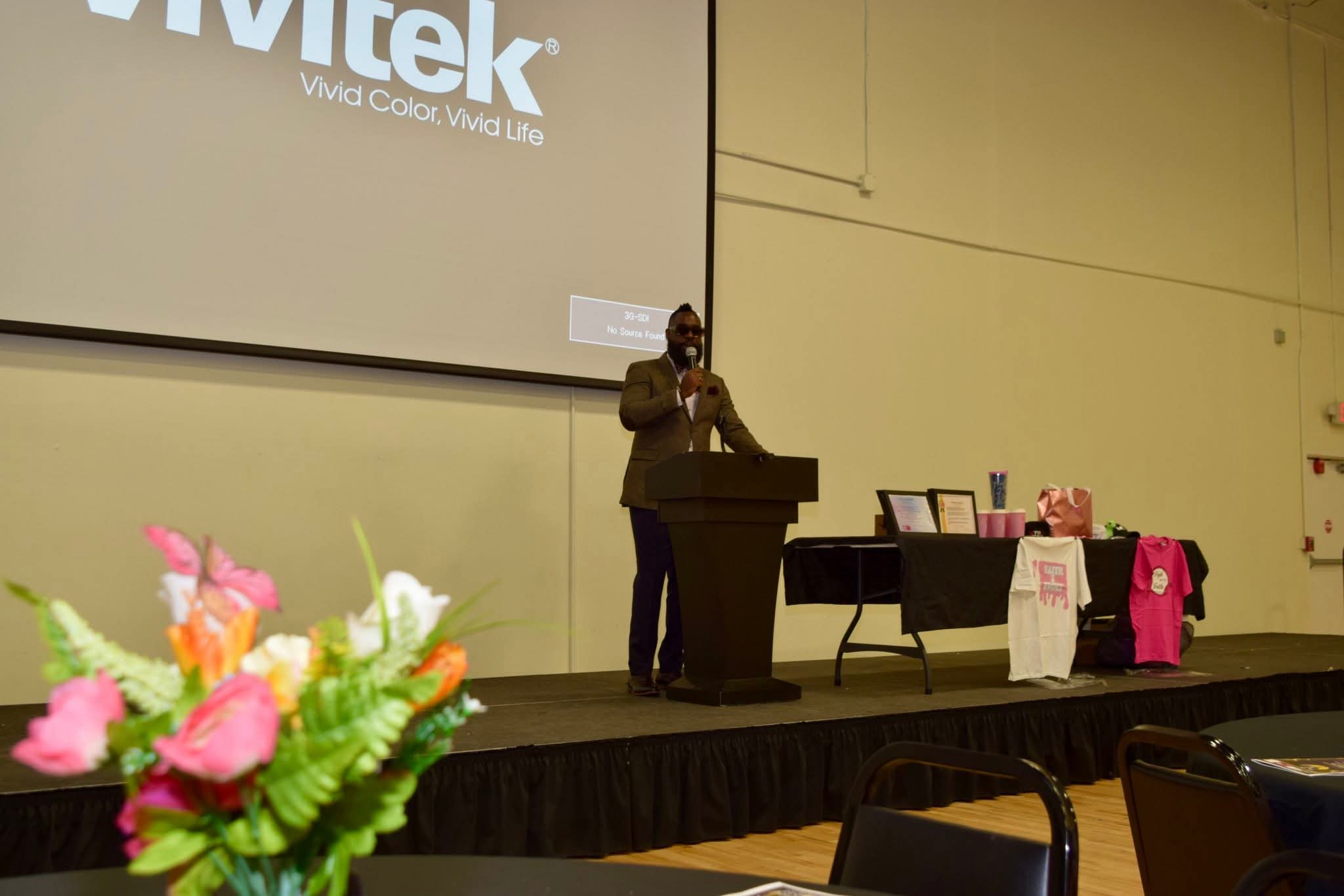 Person speaking at a podium in a large room with a presentation screen and tables with pink items, chairs in foreground.