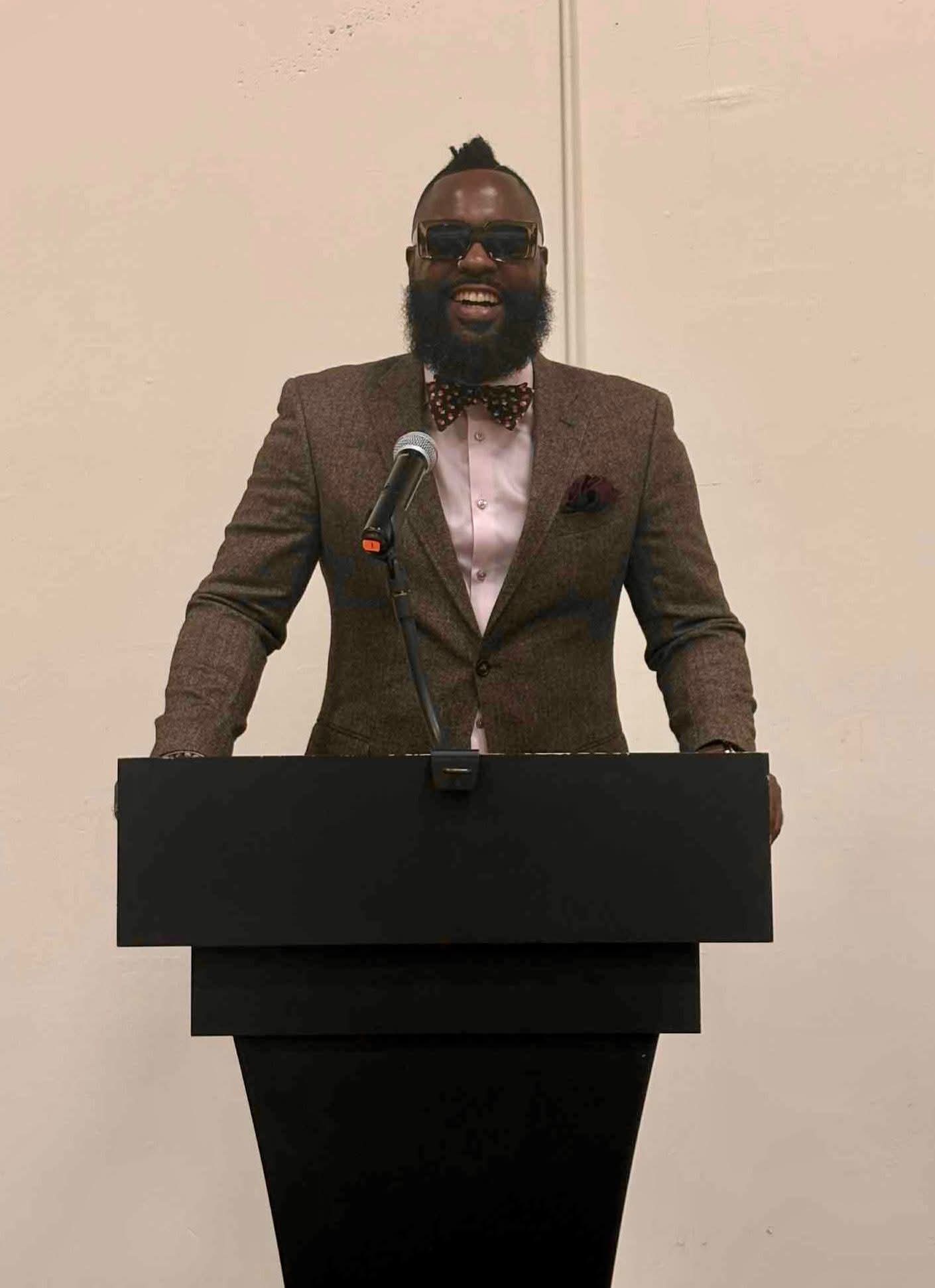 Man standing behind a podium, smiling, wearing a suit, glasses, and a bow tie, against a plain background.