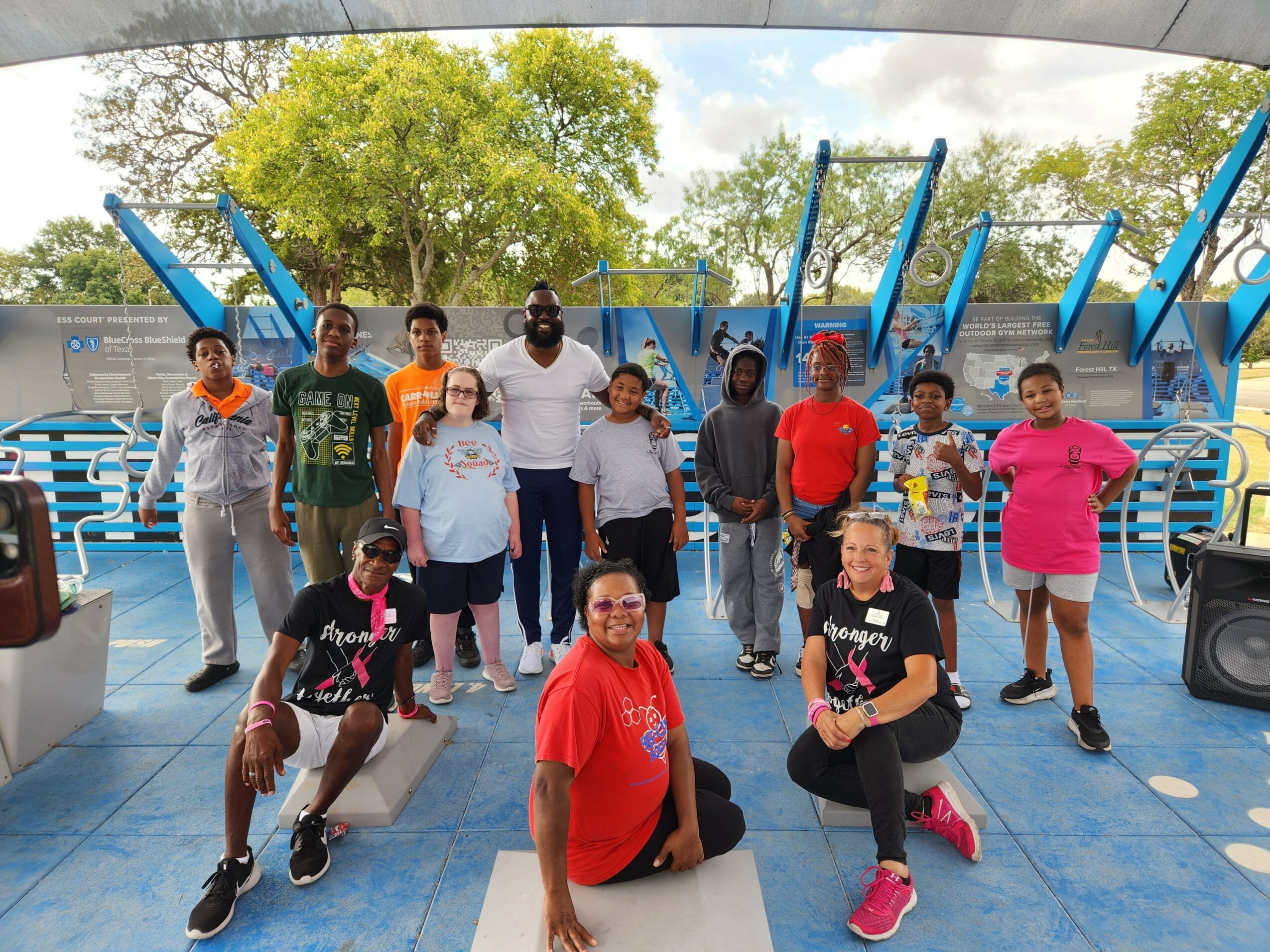 Group of children and adults on a playground with blue structures, trees, and a blue sky in the background.