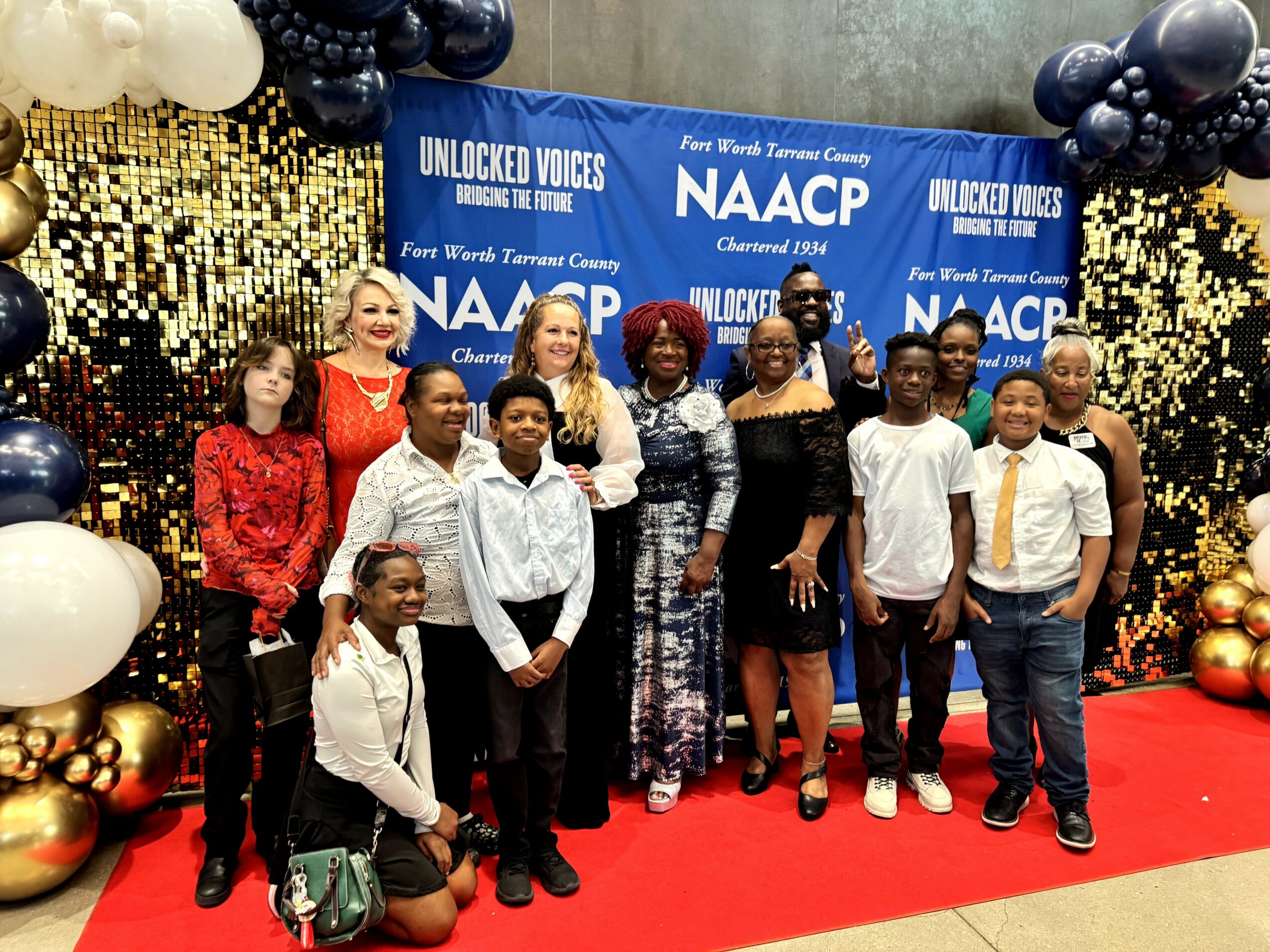 Group of people posing on a red carpet in front of a blue backdrop with gold and black balloons, celebrating an event.