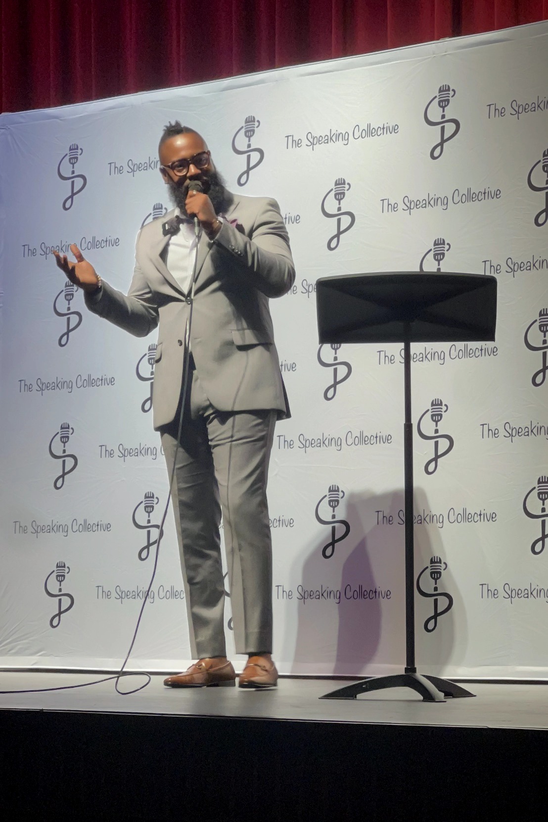 Man in gray suit speaking on stage with a microphone, standing in front of a backdrop with logos, next to a music stand.