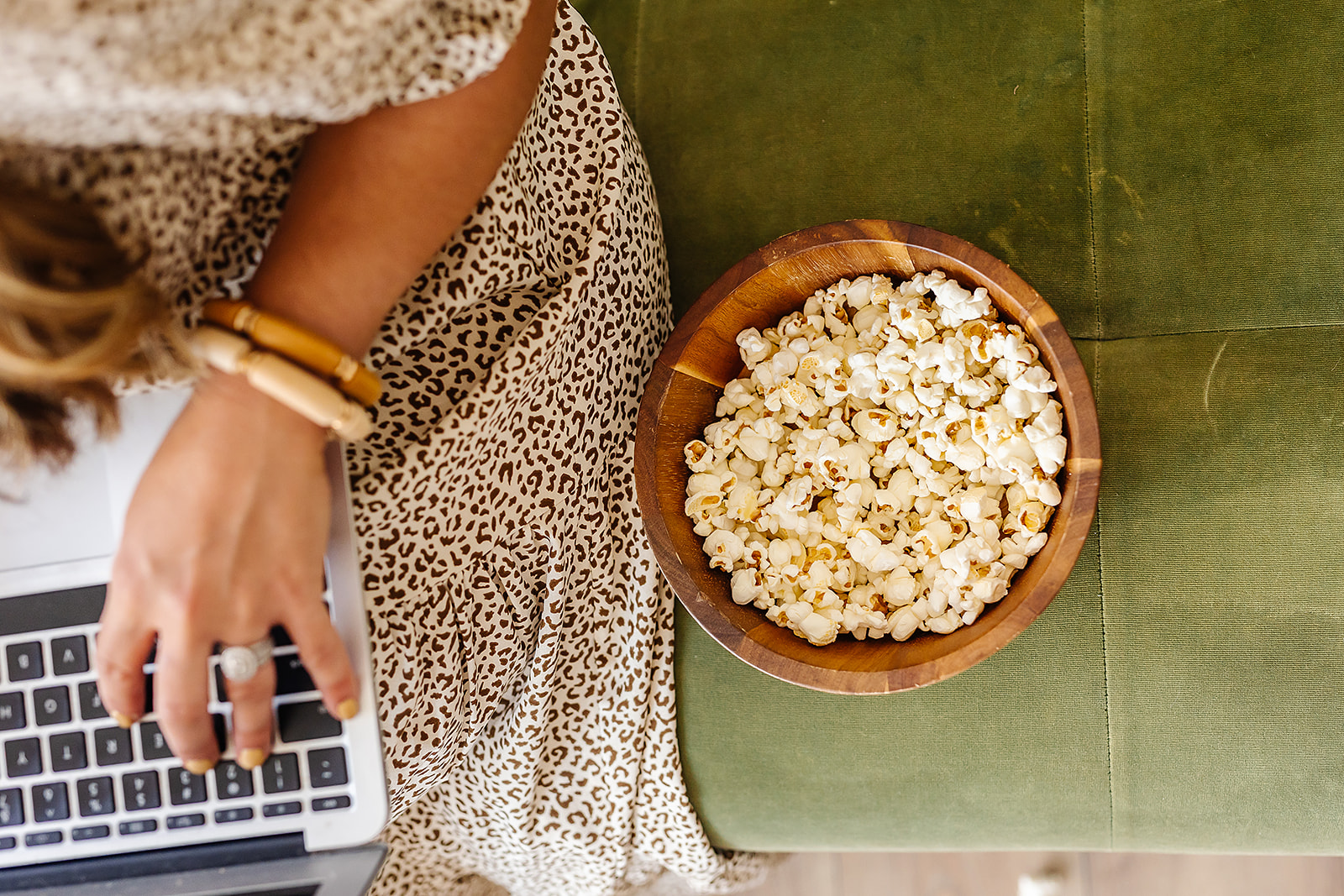 Person sitting on green surface with a bowl of popcorn nearby, using a laptop, wearing a leopard print outfit and bracelets.