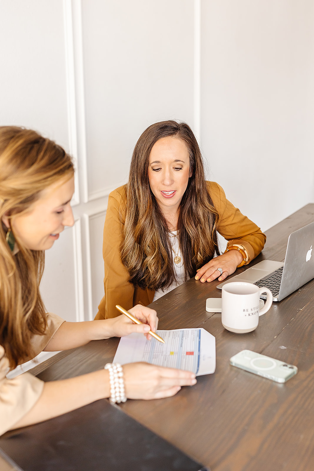 Two women sit at a table, one writing on a notepad, the other speaking, with a laptop and mug present.