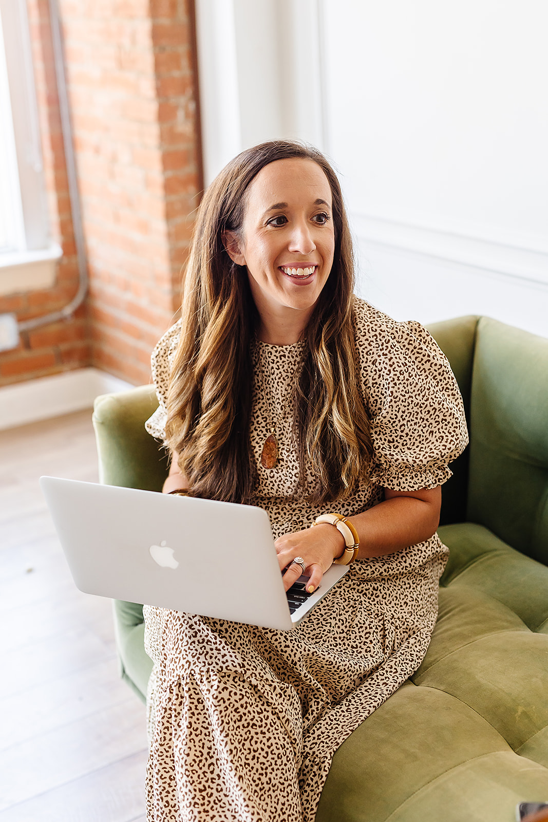 Woman with long hair smiling, sitting on a green sofa with a laptop, in a bright room with brick wall and large window.