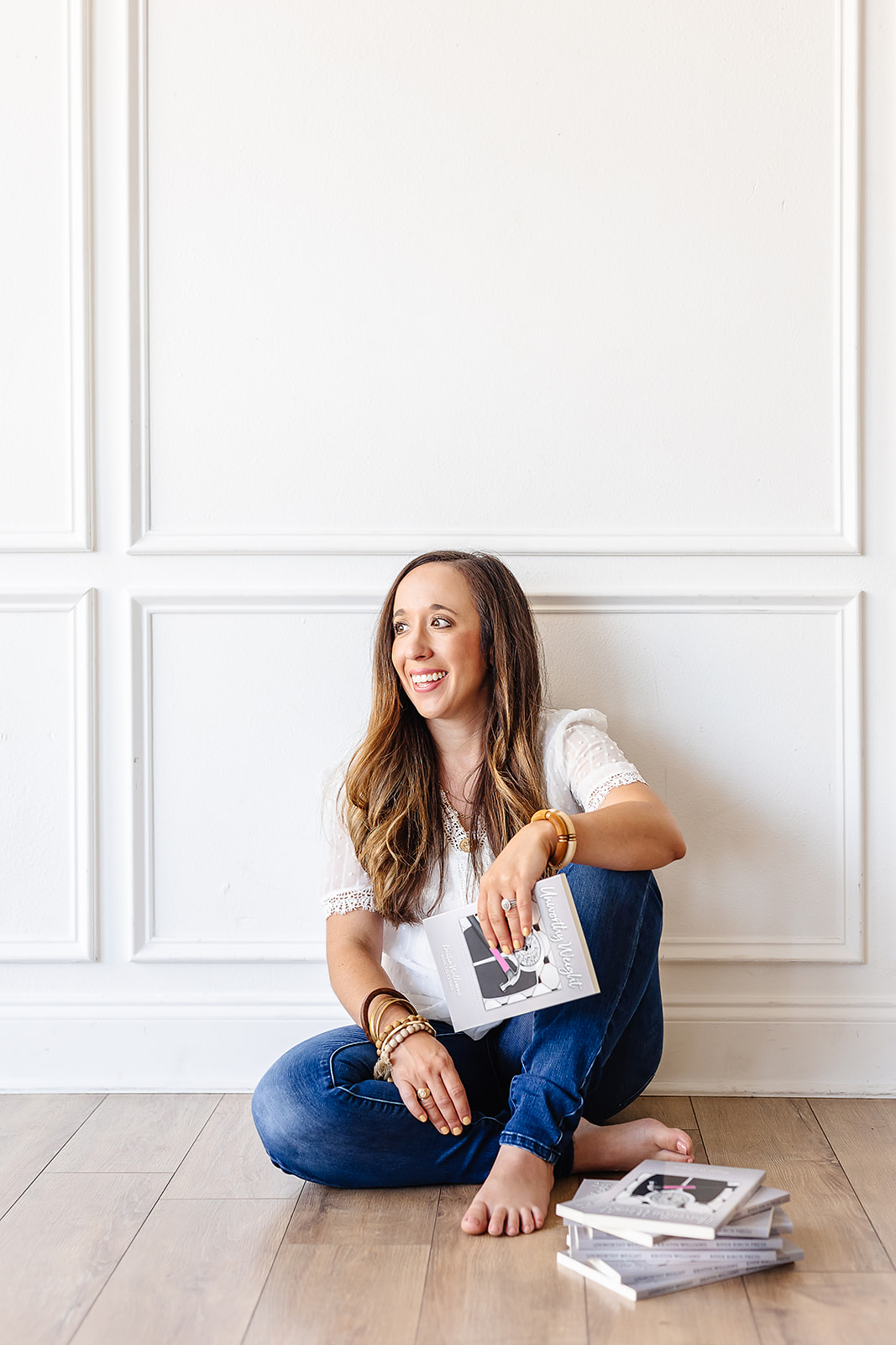 Woman sitting on floor, smiling, holding a magazine, with more magazines on the floor, against a white paneled wall.
