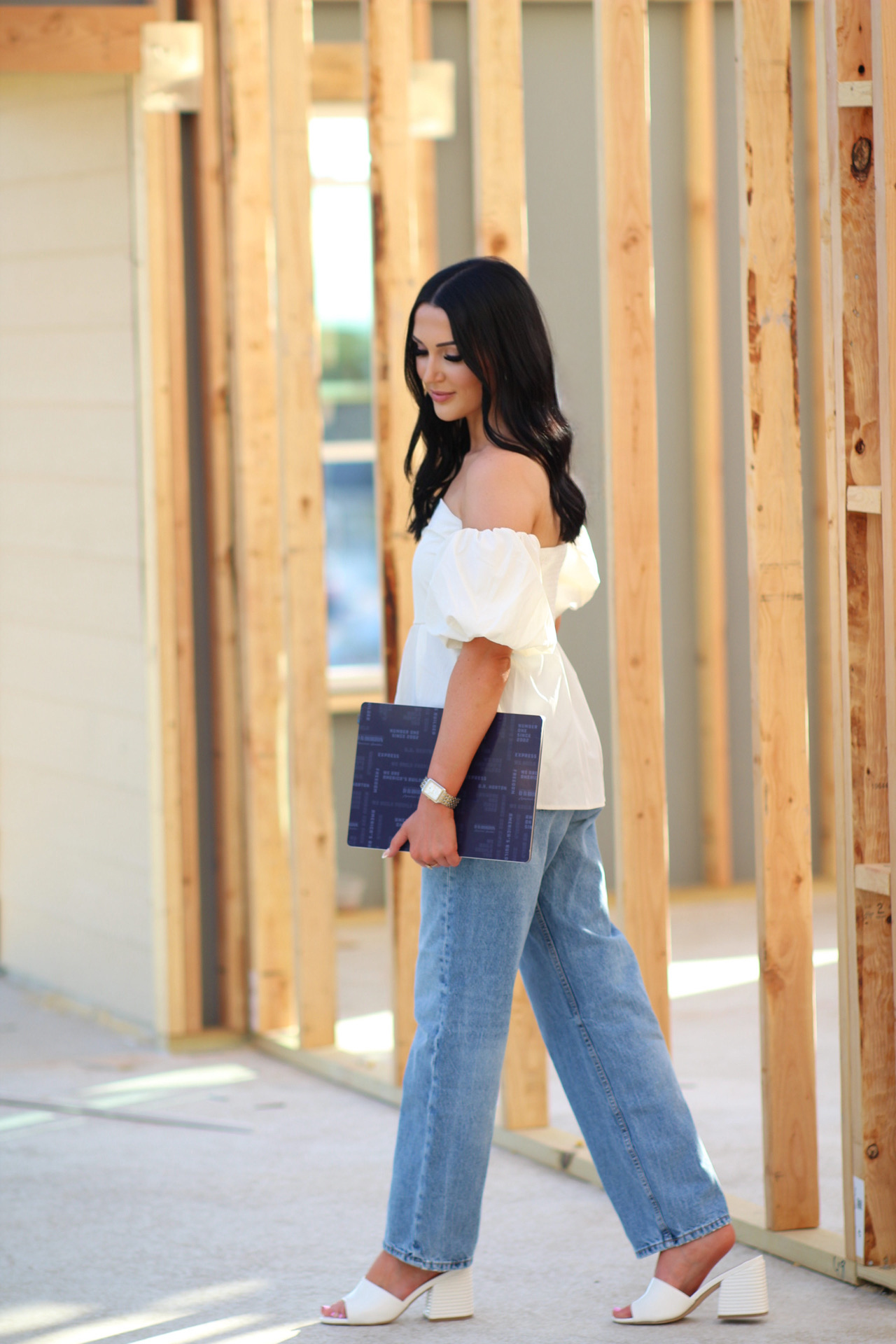 Woman with dark hair in white off-shoulder top and jeans holding a blue folder, standing among wooden framing outdoors.