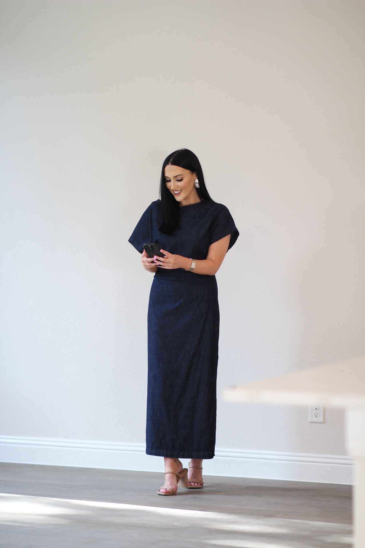 Woman standing against a plain wall, wearing a long dark dress, with hands clasped in front, looking to the side.