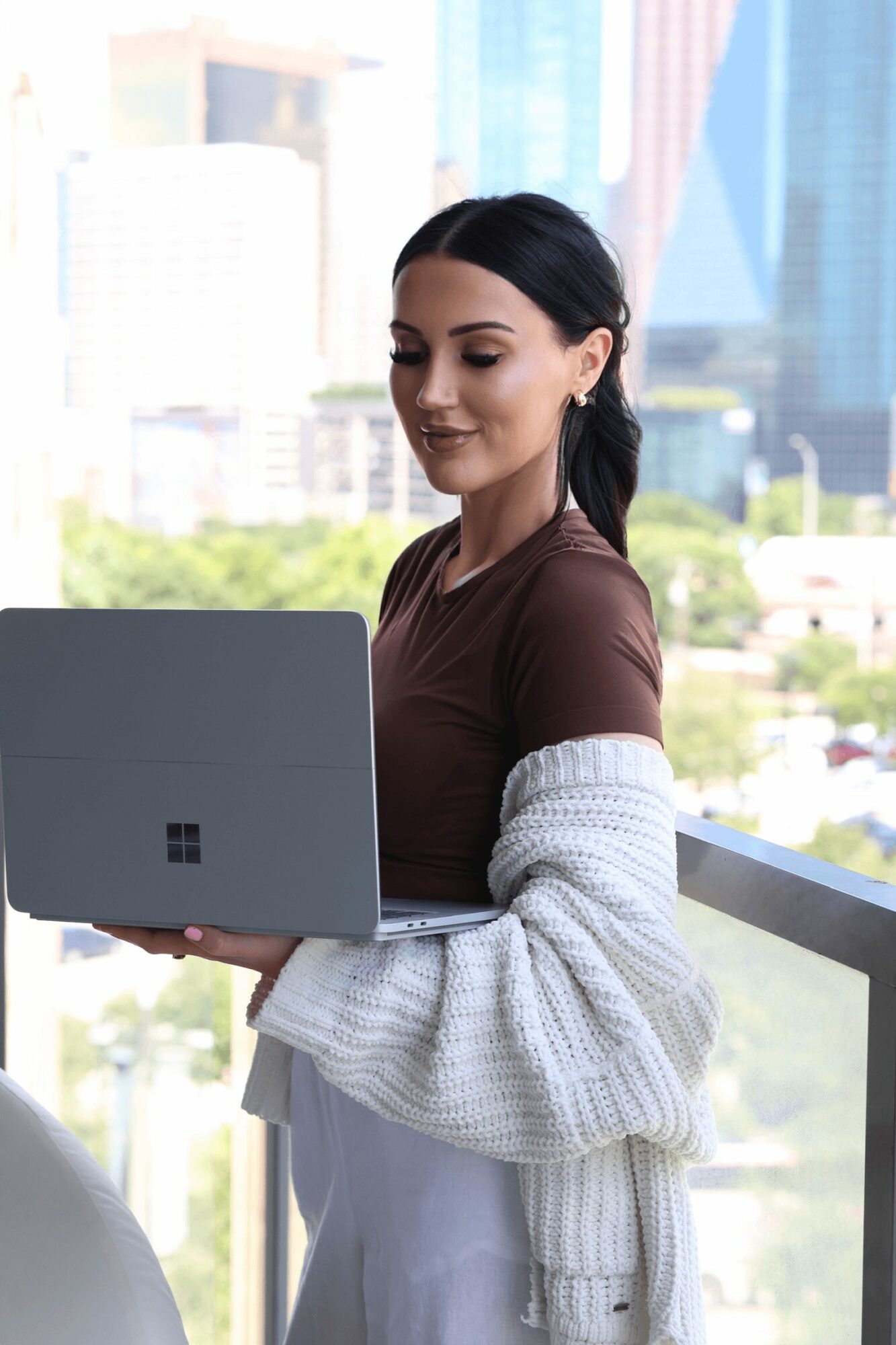 Young woman with dark hair in a ponytail using a laptop outdoors, cityscape background, wearing a brown top and white cardigan.