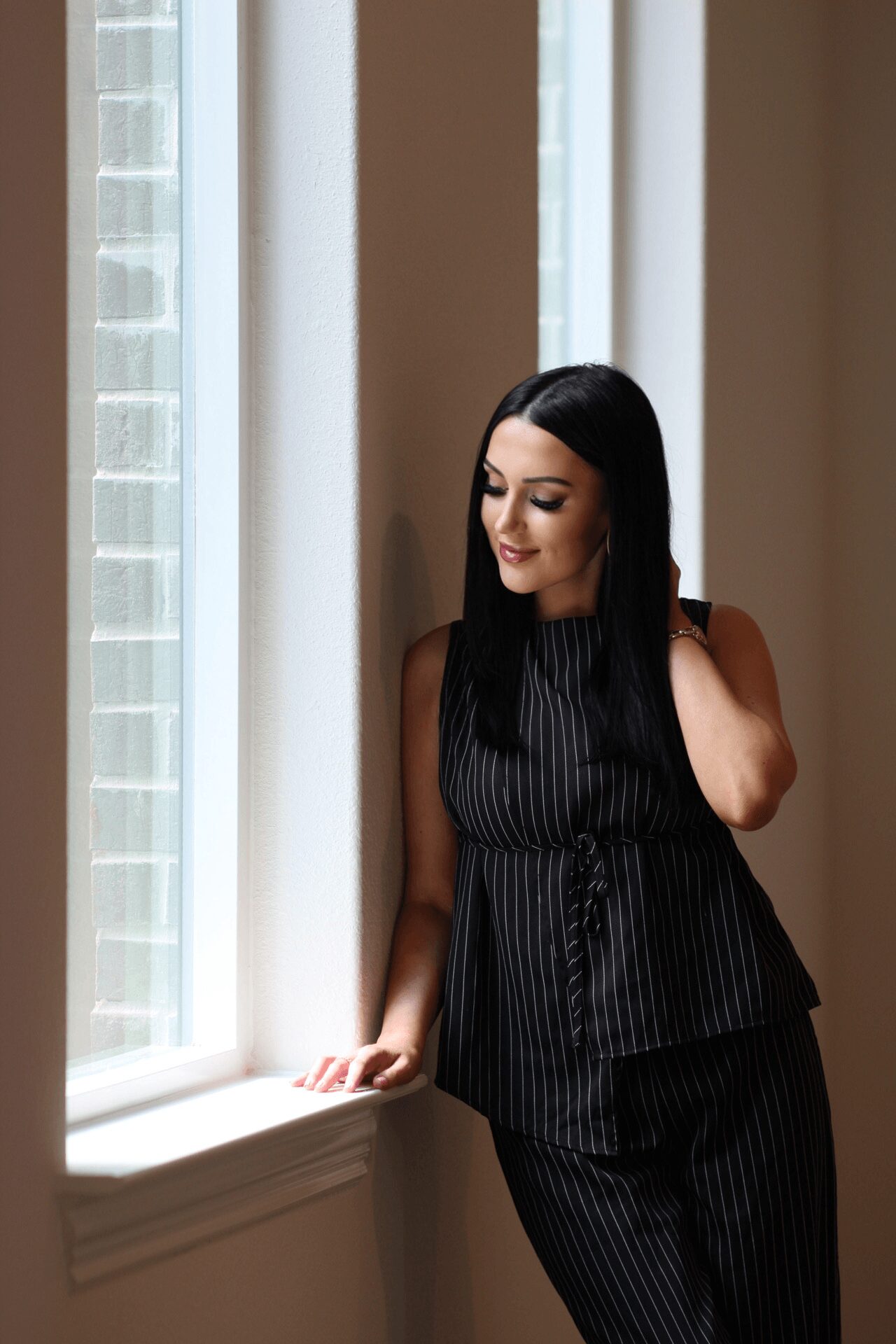 Woman with long dark hair in a black pinstripe outfit standing by a window, looking down, with natural light illuminating her.