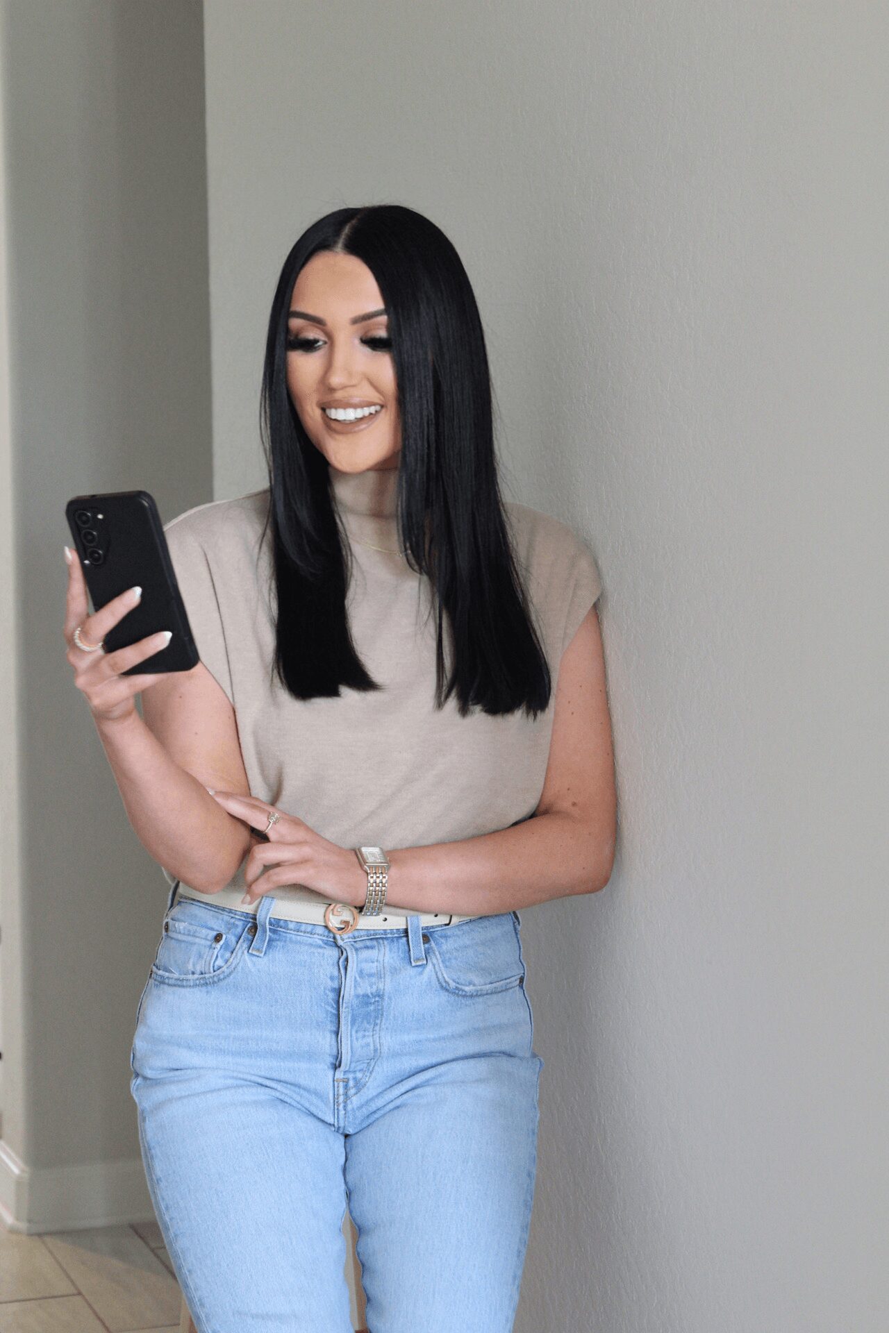 Young woman with long black hair smiling while looking at her phone, wearing a beige top and light blue jeans.