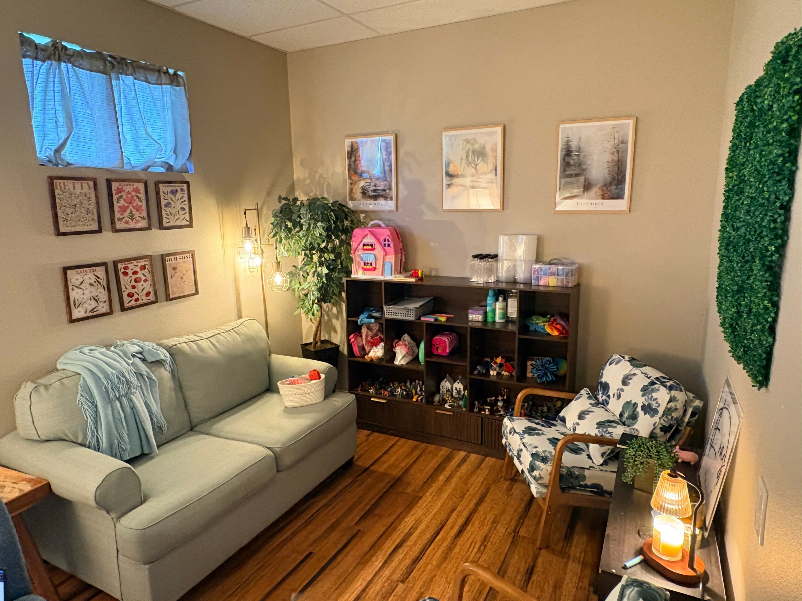 Living room with a light-colored sofa, patterned armchair, bookshelf, wall art, and a small table with a lamp.