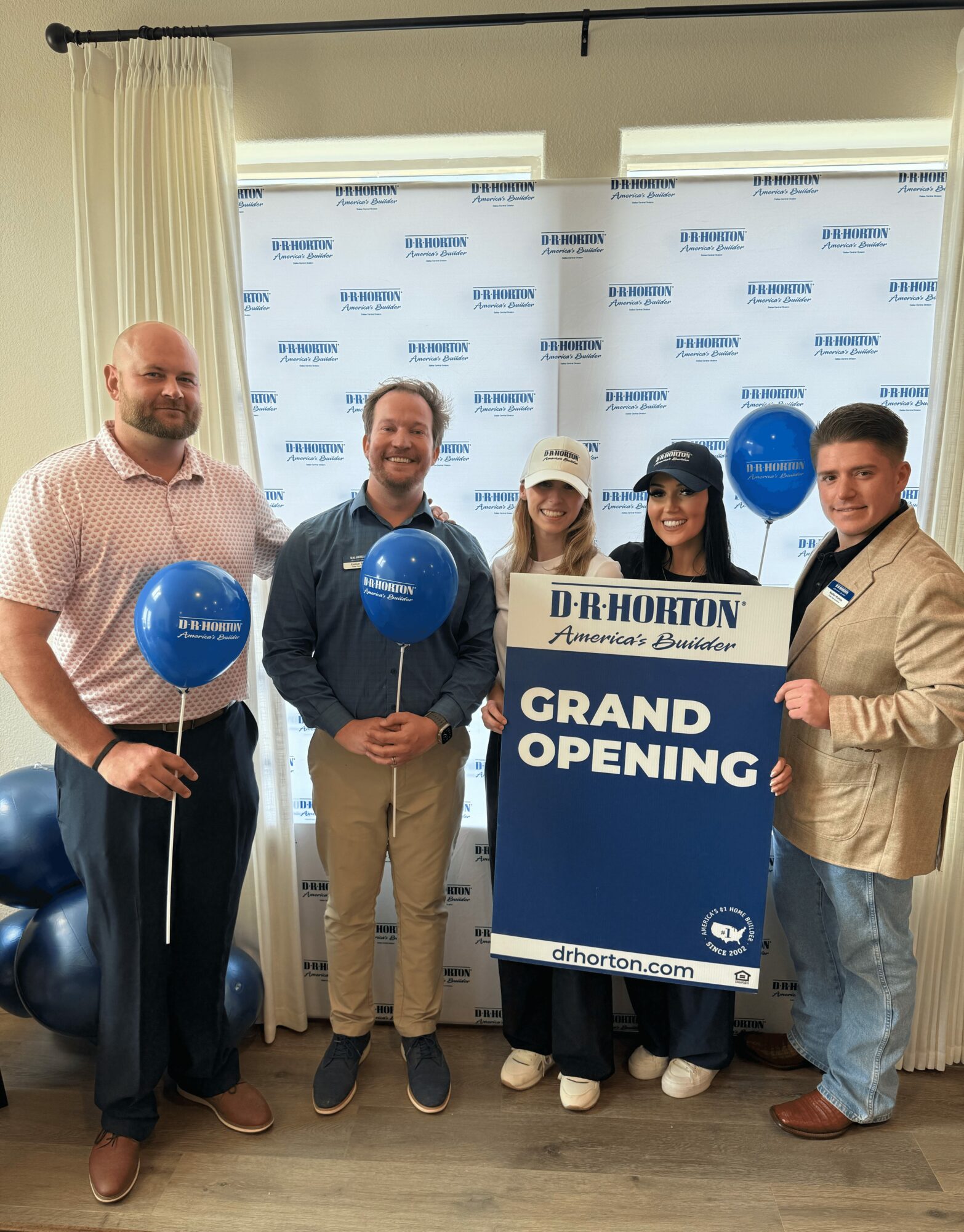 Five people standing indoors holding balloons and a sign that reads 'Grand Opening' in front of a backdrop with text. They are smiling.