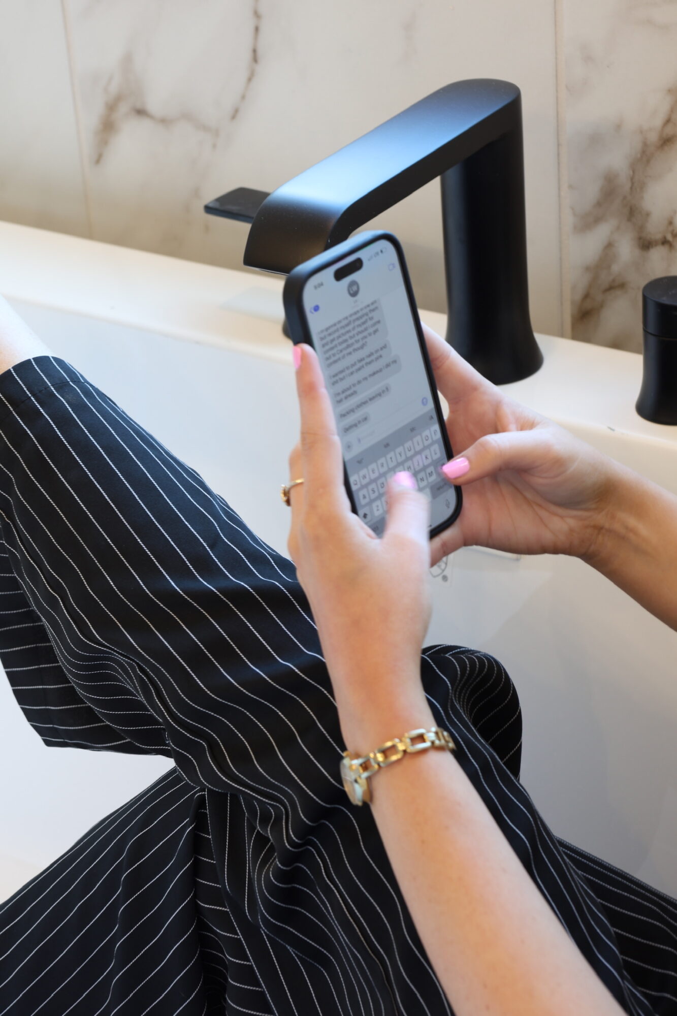 Person holding a smartphone over a sink with a black faucet, wearing a striped outfit and a gold bracelet.