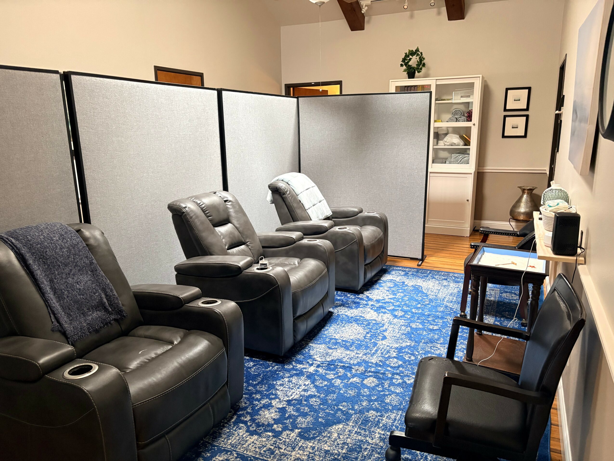 Room with three black recliner chairs, a blue patterned rug, and a white cabinet with glass doors, with a divider in the background.