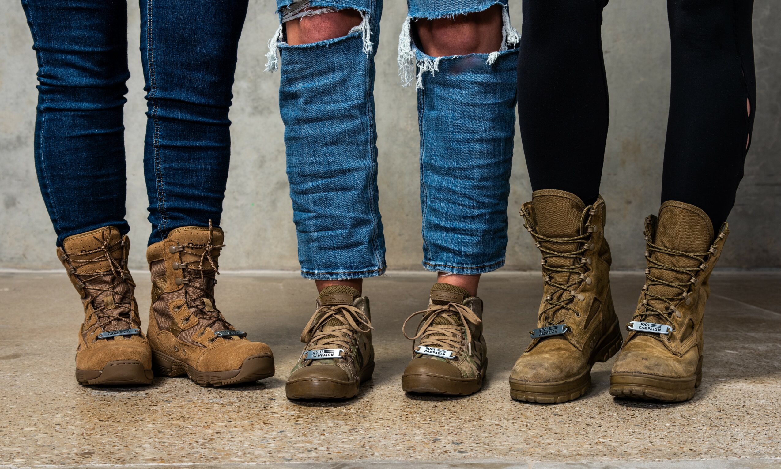 Three people wearing jeans and boots standing on a concrete floor, with a gray wall background.