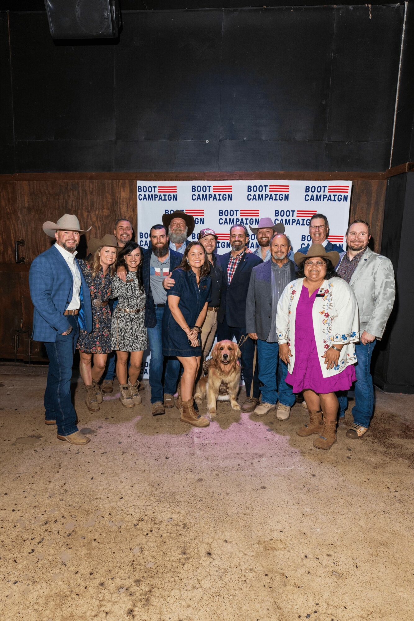 Group of people, including a dog, standing on stage with a campaign backdrop, in a dimly lit room.