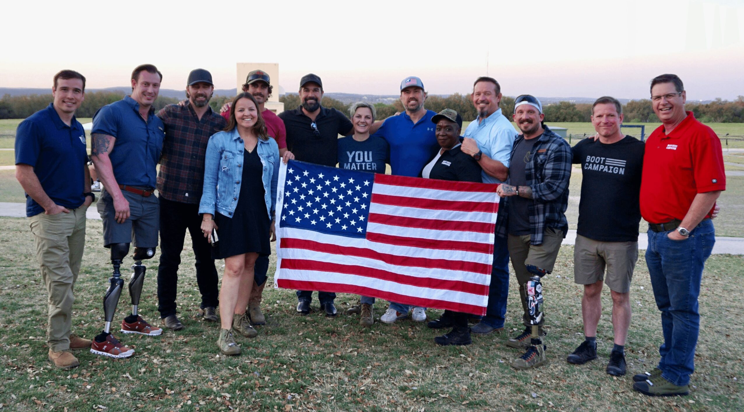 Group of people outdoors holding an American flag, standing on grass with a cloudy sky background.