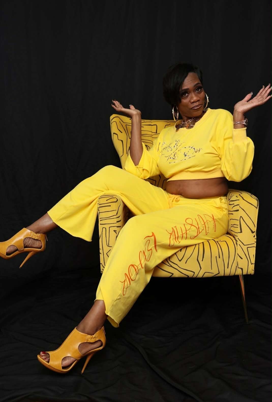 Woman in yellow outfit sitting on a patterned chair with arms raised, black background, wearing high heels.