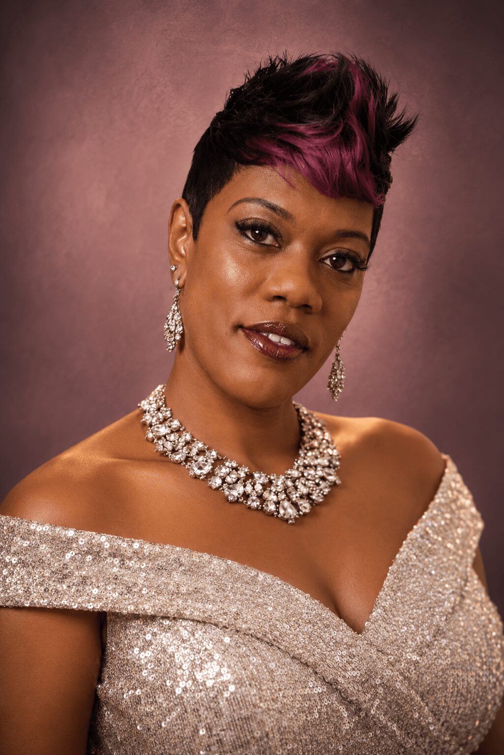 Woman with short hairstyle, wearing a sparkly off-shoulder dress and jewelry, against a plain background.