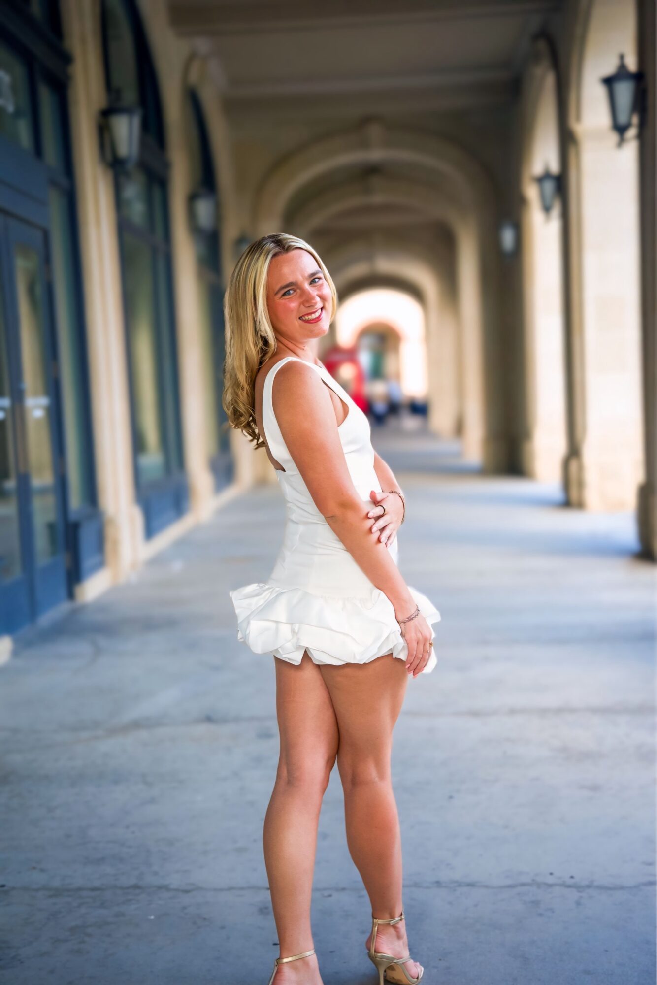 Young woman in white dress standing in a covered walkway with arches, smiling, with her arms crossed, sunlight illuminating her.