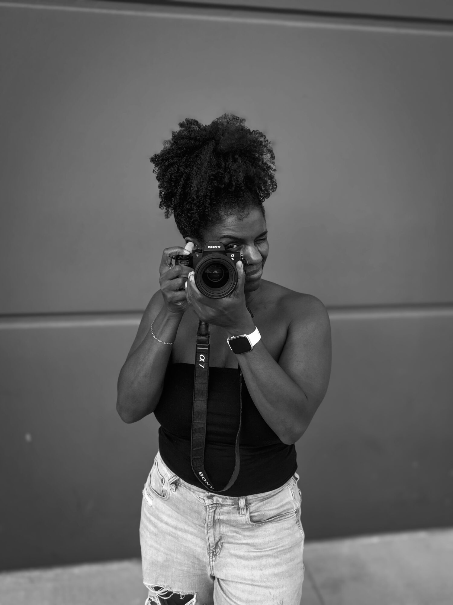 Woman with curly hair taking a photo with a camera, wearing a black top and light-colored shorts, standing against a plain wall.