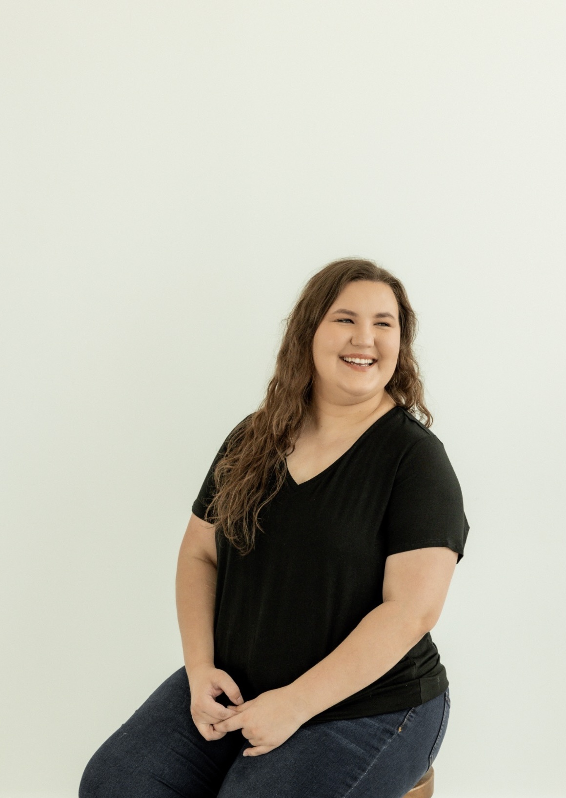 Smiling woman with long wavy hair wearing a black shirt and jeans, standing against a plain light background.