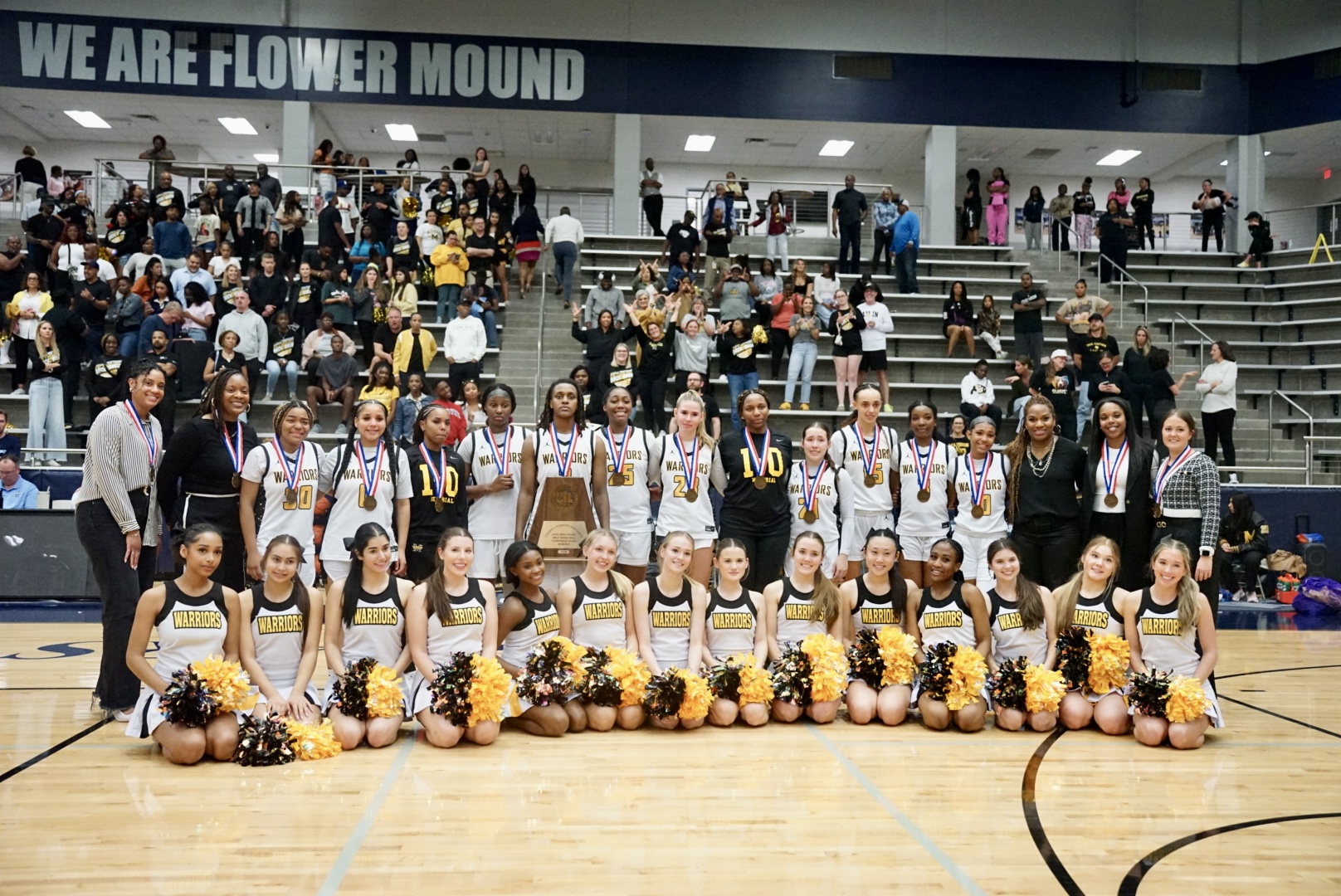 Cheerleading team in uniform with pom-poms, posing on basketball court, with spectators in stands behind them.