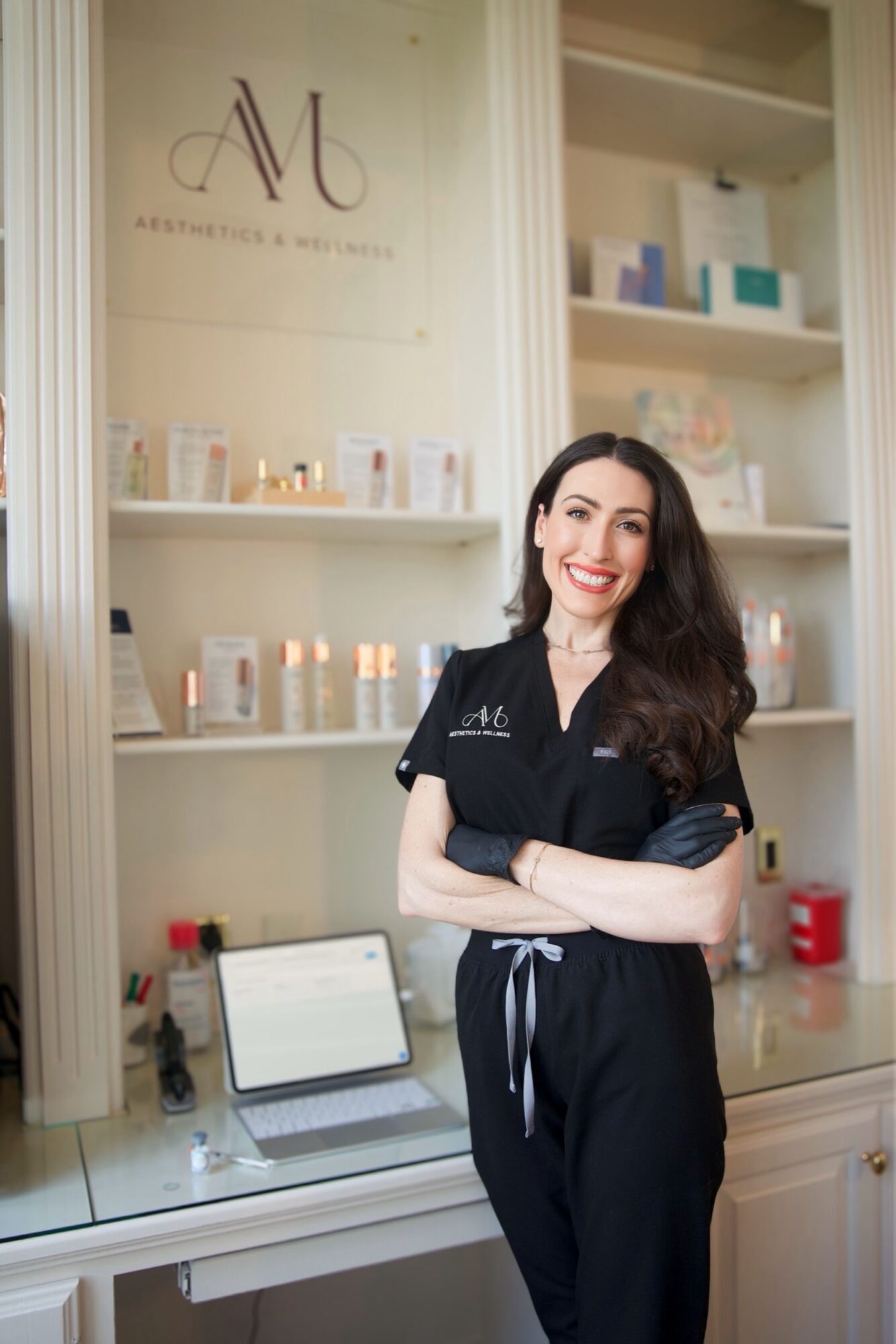 Smiling woman with long dark hair in black scrubs standing in a skincare clinic, with shelves of products behind her.