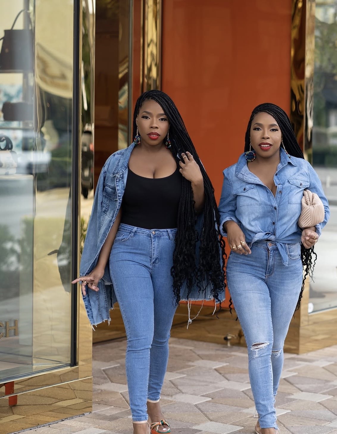 Two women walk together indoors, both wearing denim jackets and jeans, one holding a purse, with a reflective wall behind them.