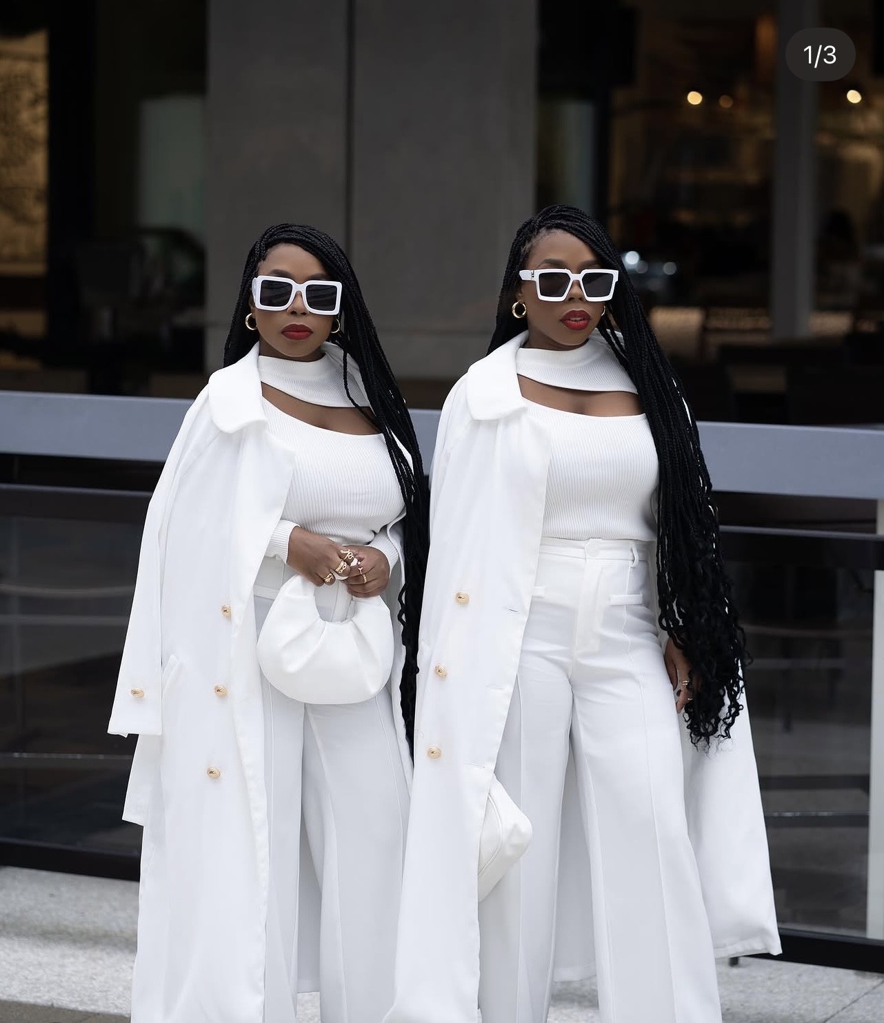Two women in white outfits with large sunglasses stand outdoors in front of a building with glass windows.