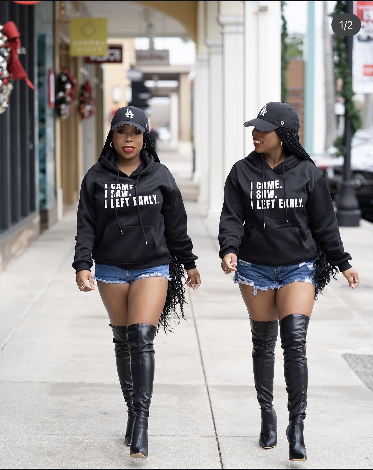 Two women walk side by side on a sidewalk, wearing black hoodies, denim shorts, and thigh-high black boots, with buildings in the background.