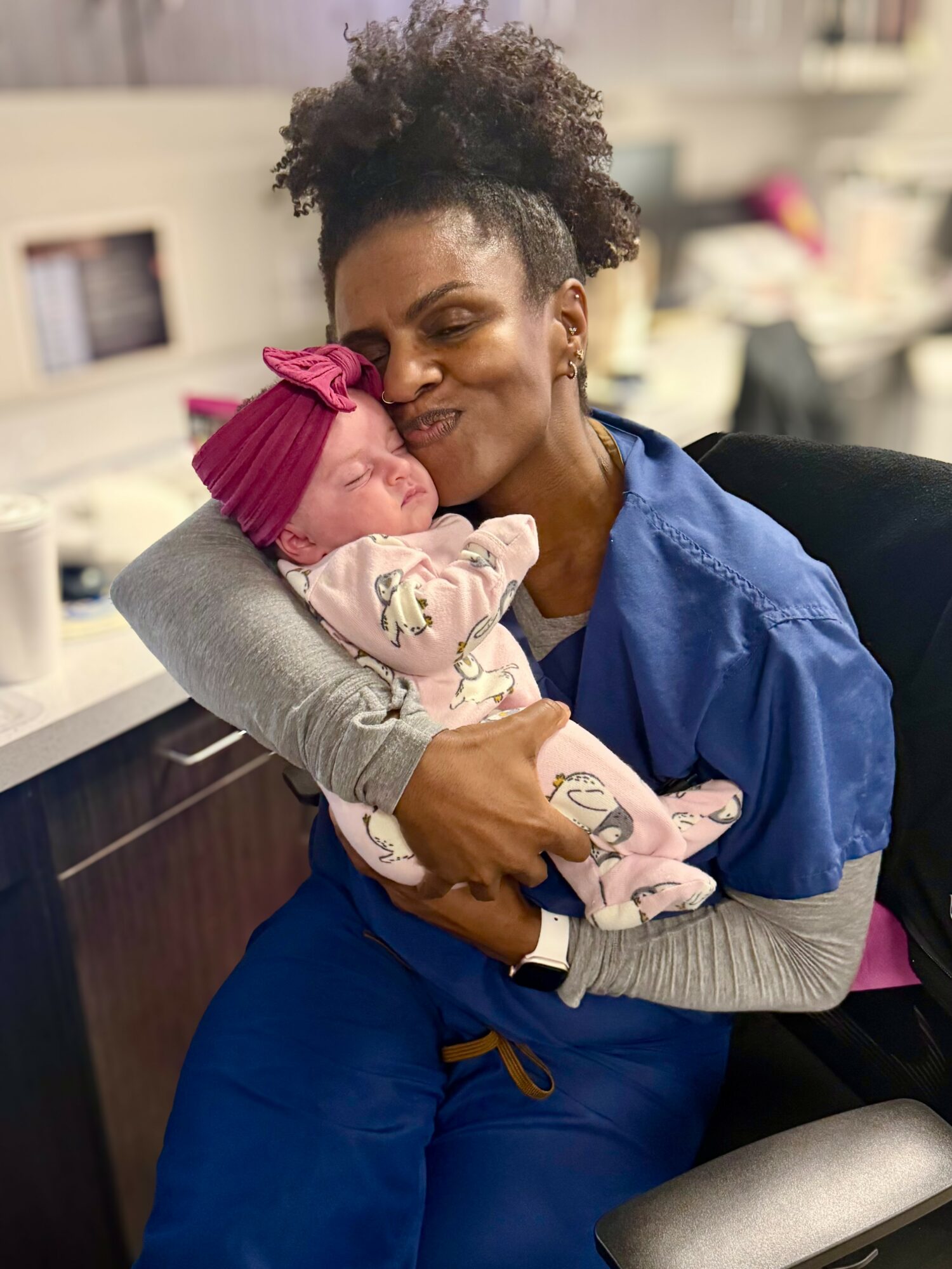 Woman holding a sleeping baby with a pink headband, smiling in a room with shelves and objects in the background.