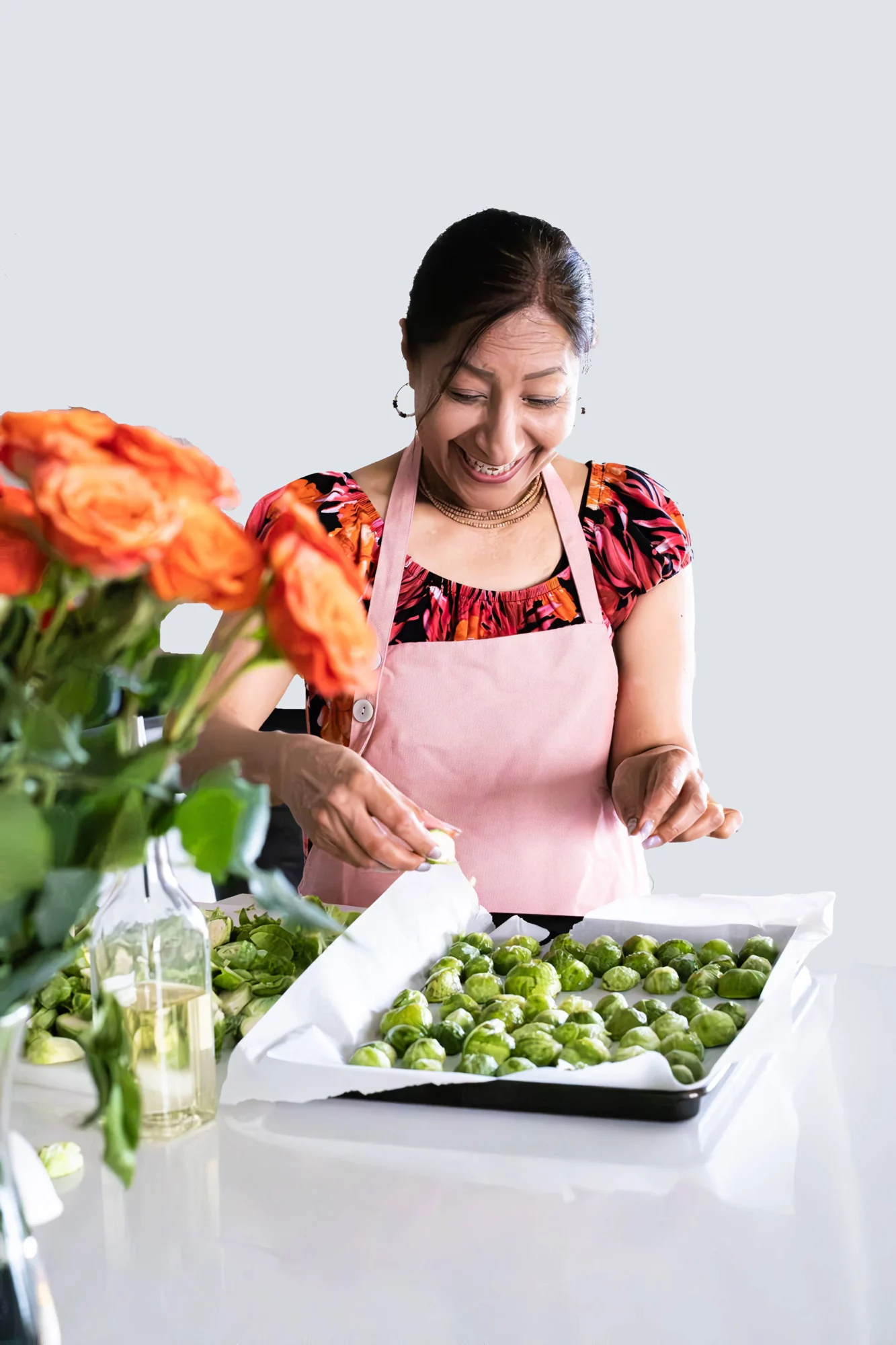 Woman smiling while harvesting green vegetables in a kitchen, wearing an apron, with flowers on the side.