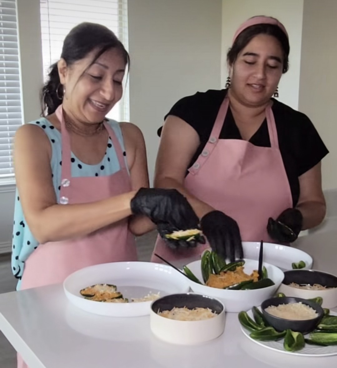 Two women wearing pink aprons preparing food in a kitchen, smiling and handling vegetables.