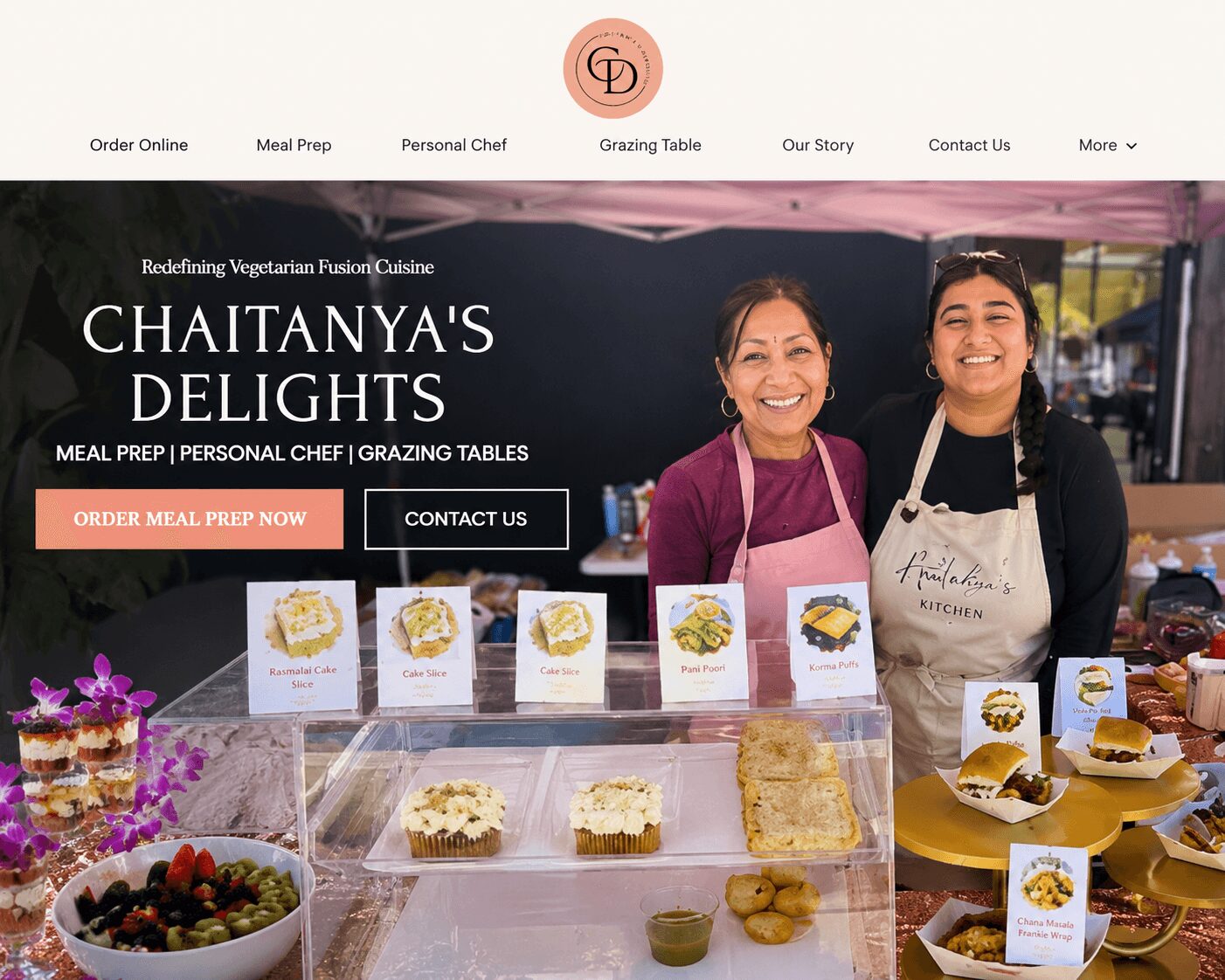 Two women smiling behind a food stall with desserts and signs, outdoor setting, pink canopy in background.