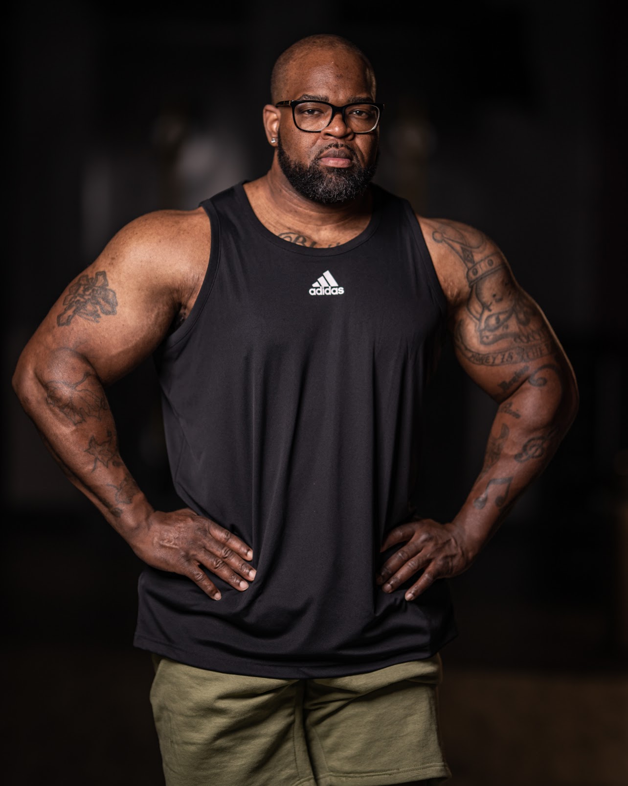 Man with glasses, beard, muscular arms, wearing a black sleeveless athletic shirt, standing with hands on hips in a dark setting.