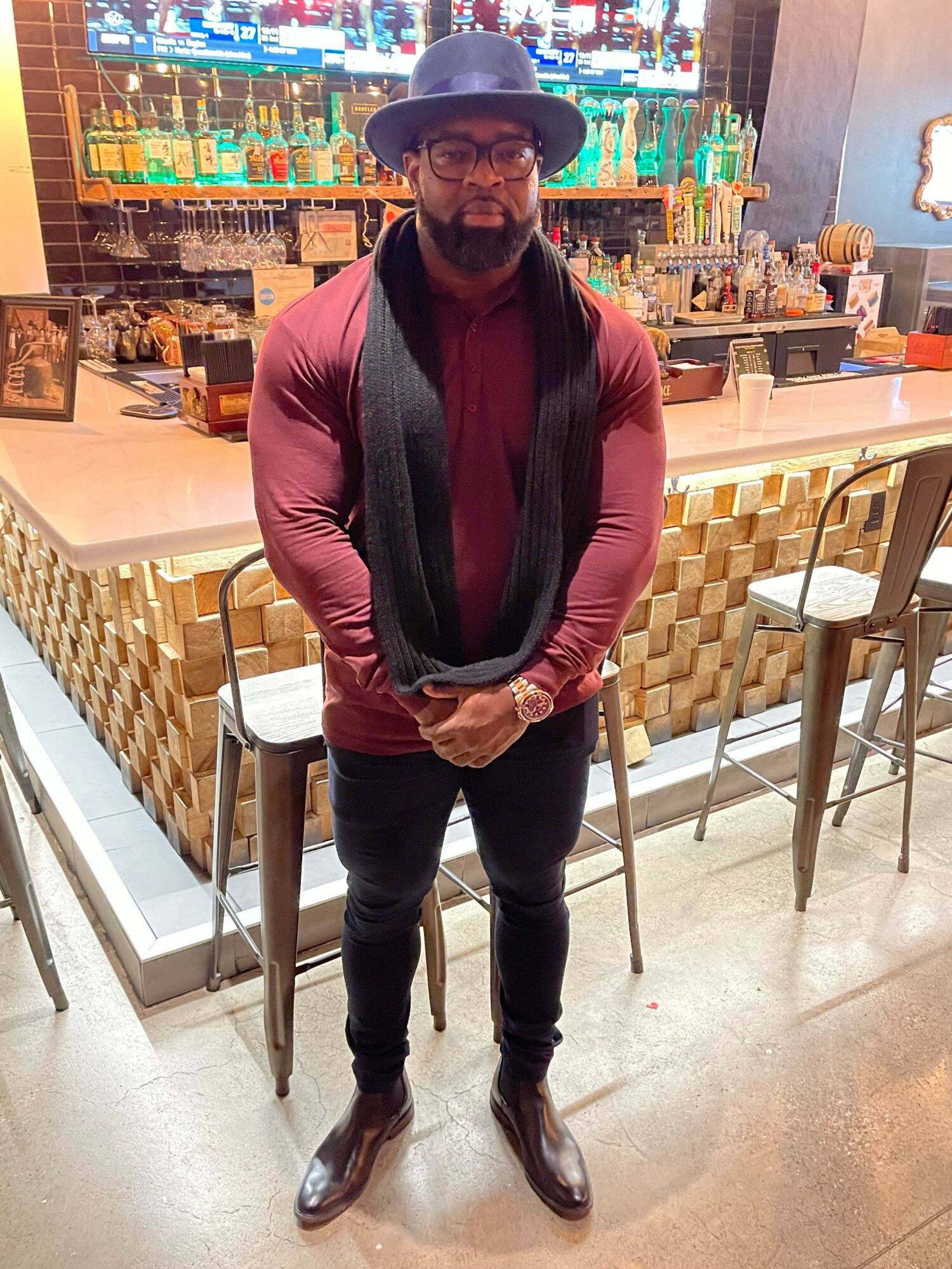 Person wearing a hat, glasses, maroon jacket, black pants, and shoes, standing indoors near a bar area.