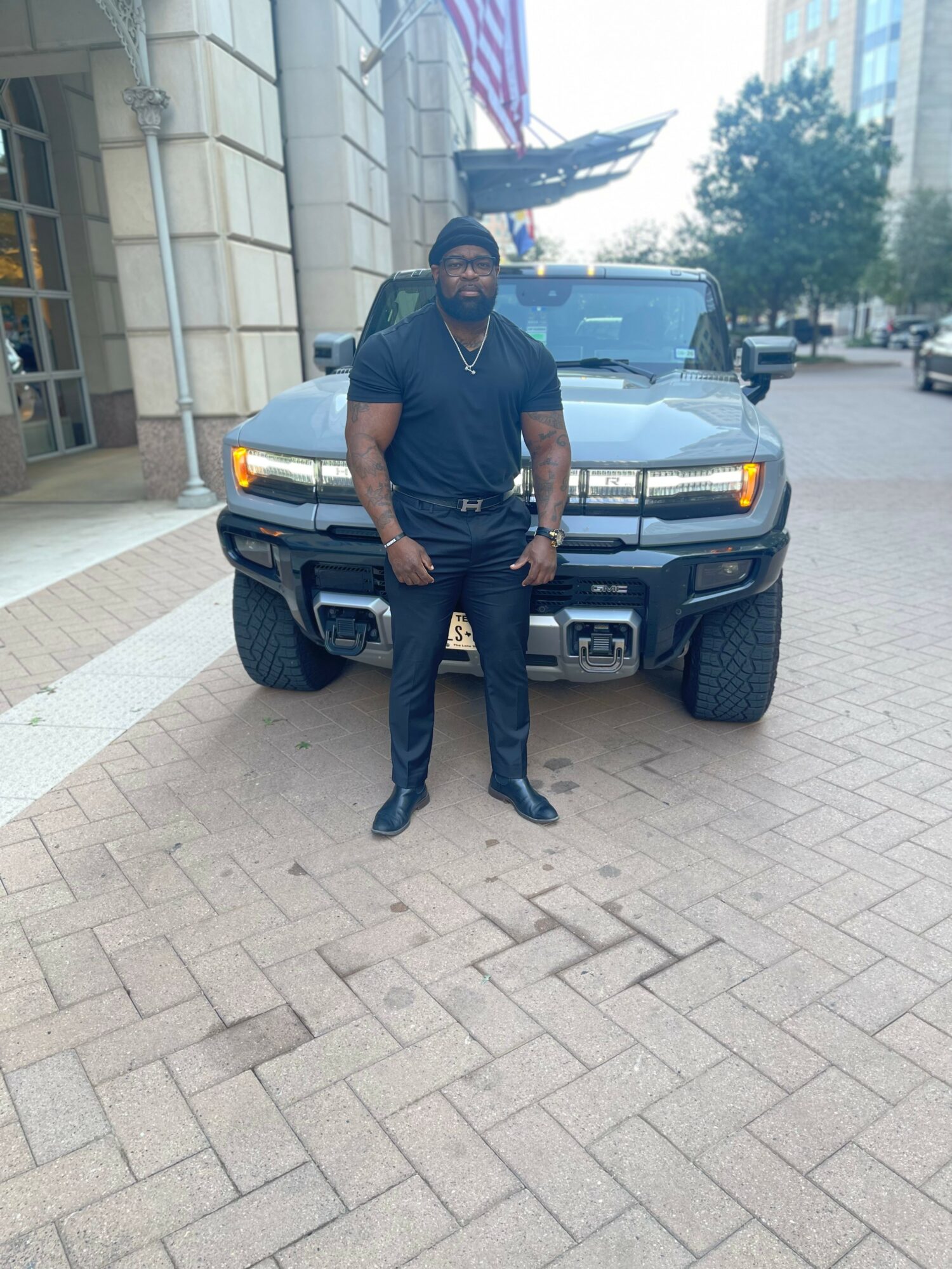 Man in black clothing standing in front of a black SUV on a city street, buildings and trees in background.
