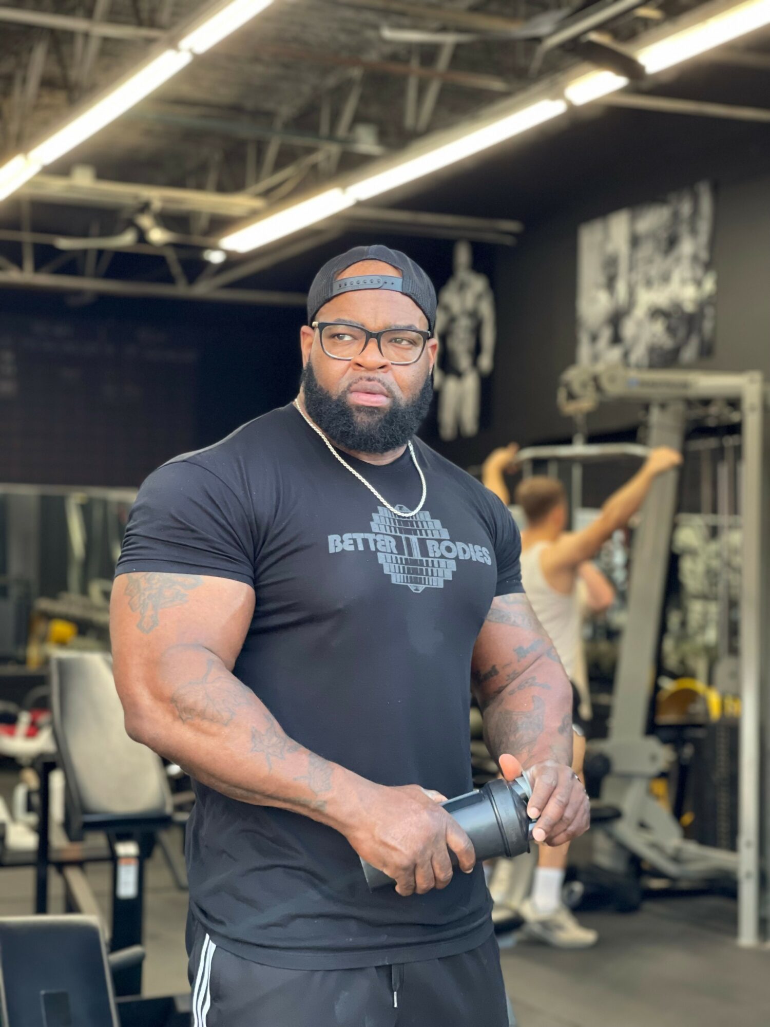 Man with glasses and a cap holding a dumbbell in a gym, with a woman exercising in the background.