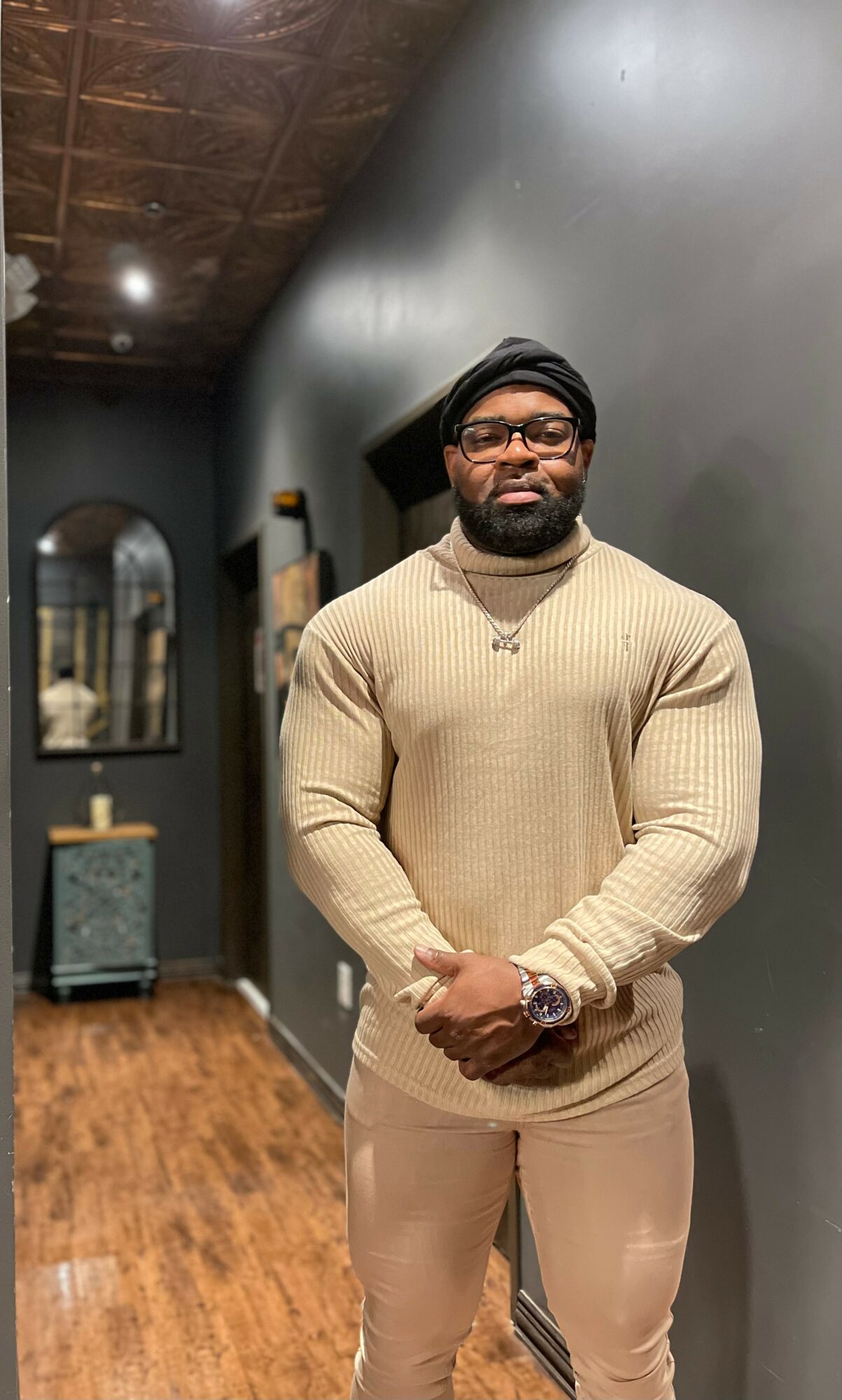 Man with glasses and a beard wearing a beige sweater and pants, standing indoors against a dark wall.