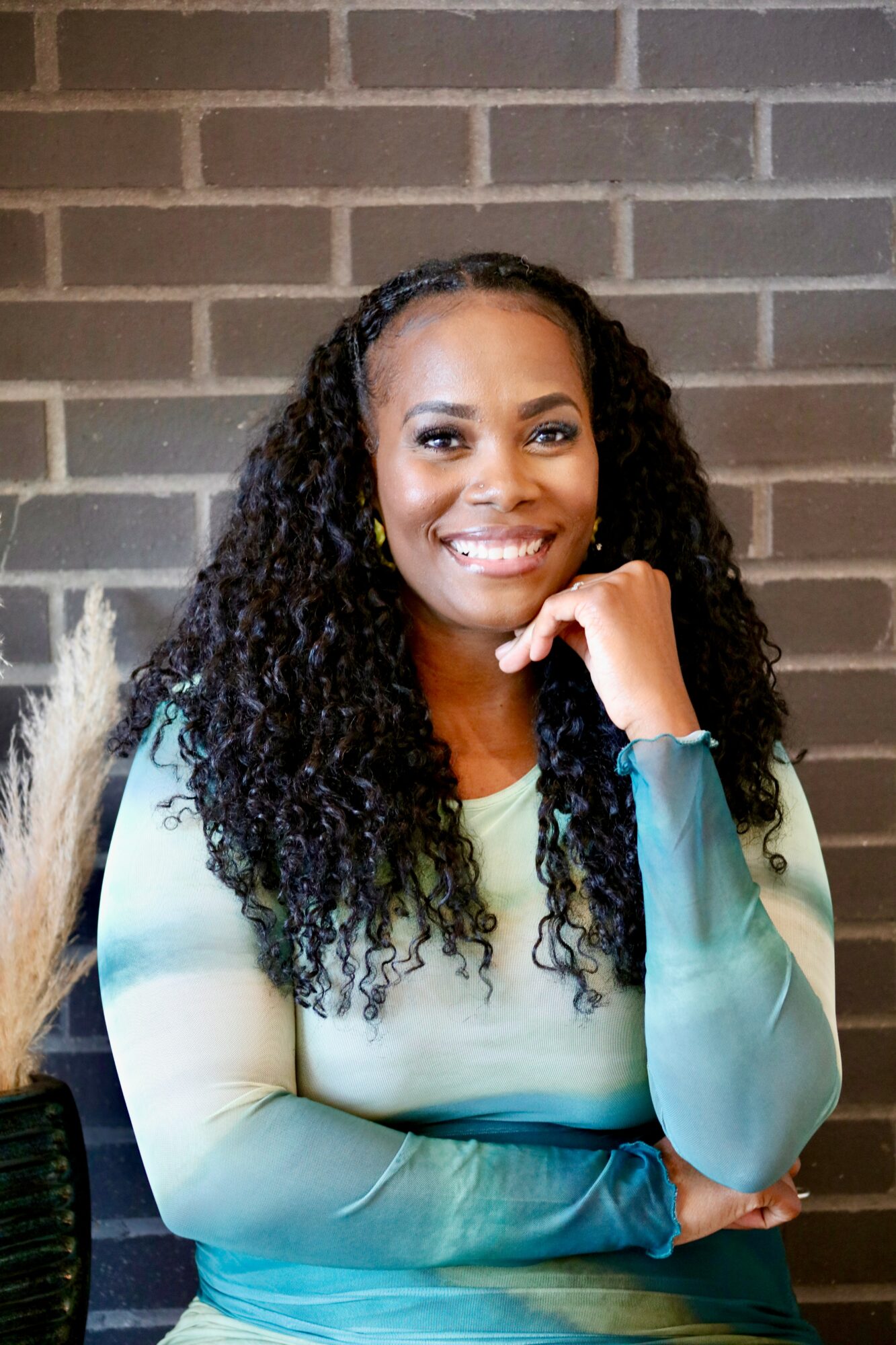 Smiling woman with curly hair, wearing a light-colored top, sitting against a brick wall, resting her chin on her hand.