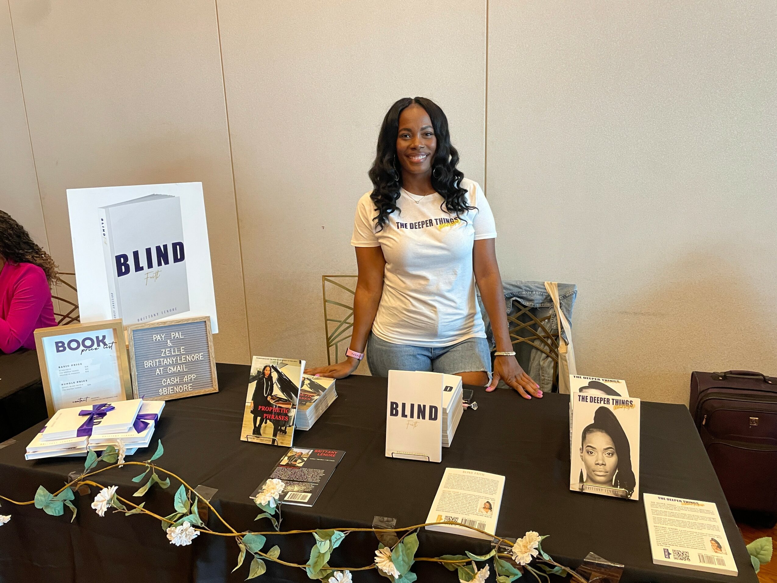 Woman standing behind a table with books and promotional materials, smiling, in an indoor setting.