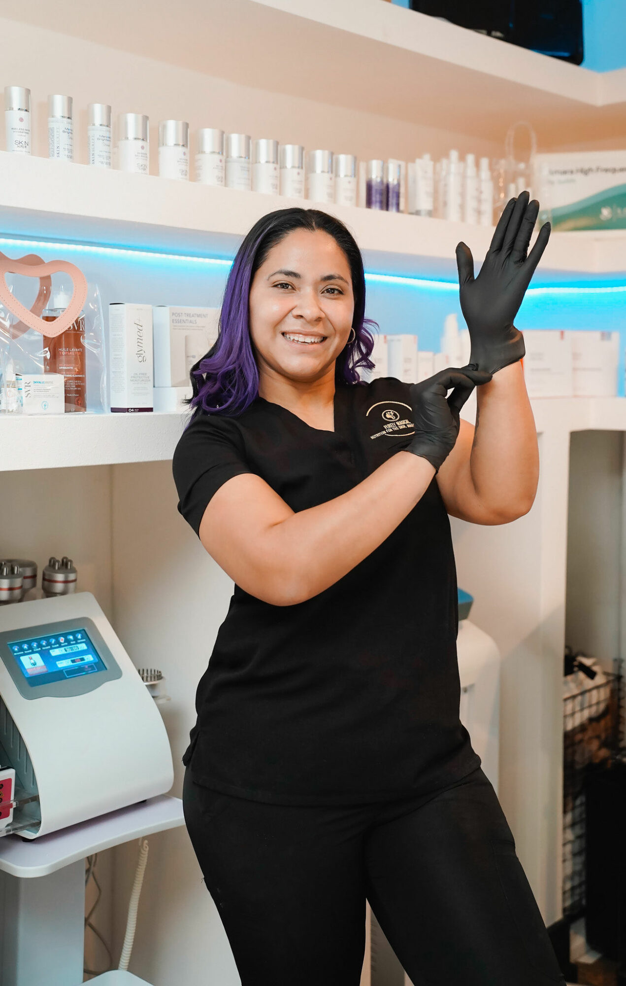 Smiling woman in black uniform putting on a black glove in a nail salon, shelves with nail polish bottles behind her.