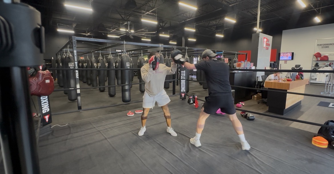 Two people sparring in a boxing gym with punching bags and equipment around them.