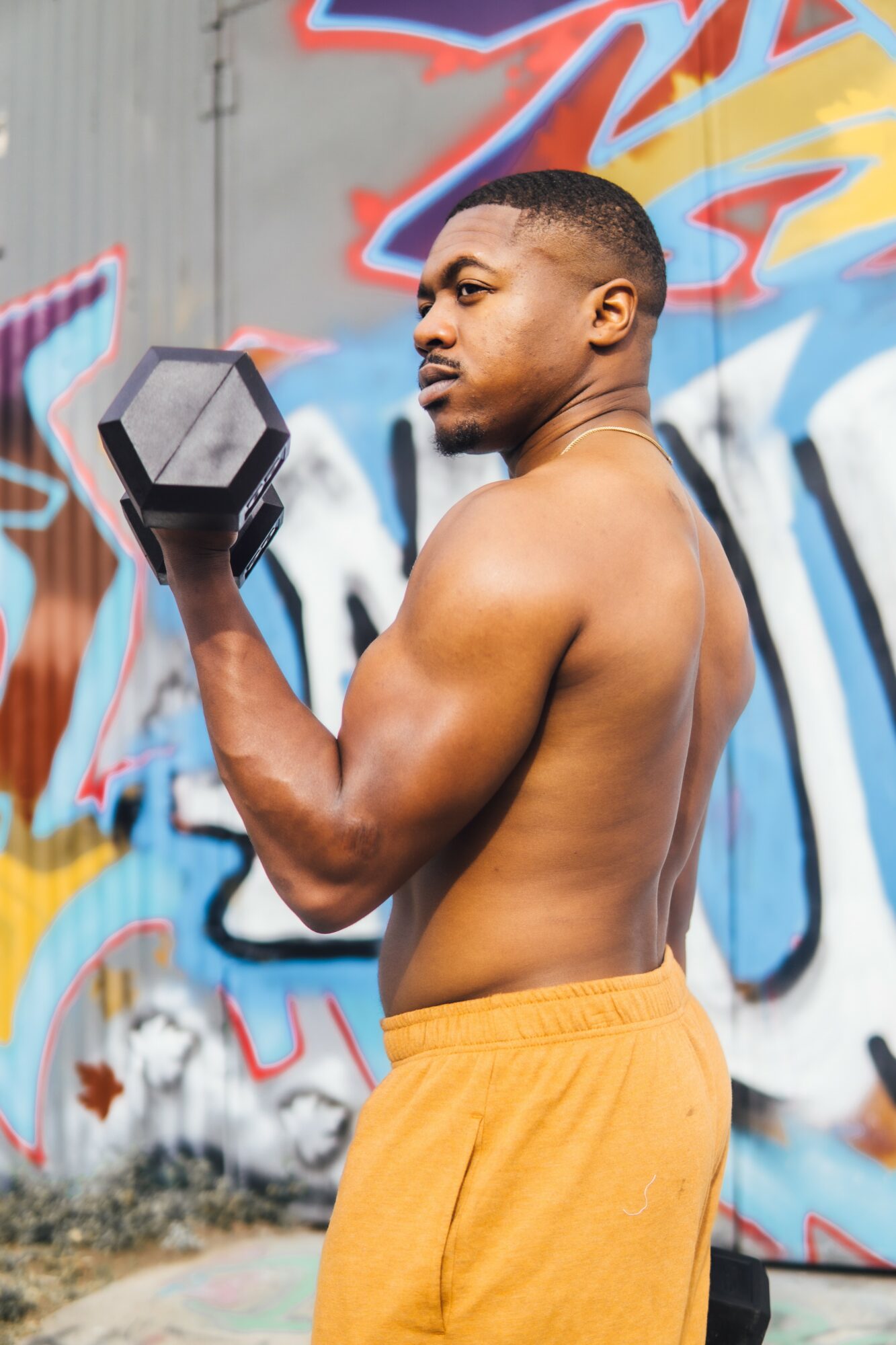 A shirtless man with muscular arms and a serious expression holds a black dumbbell against a colorful graffiti wall.