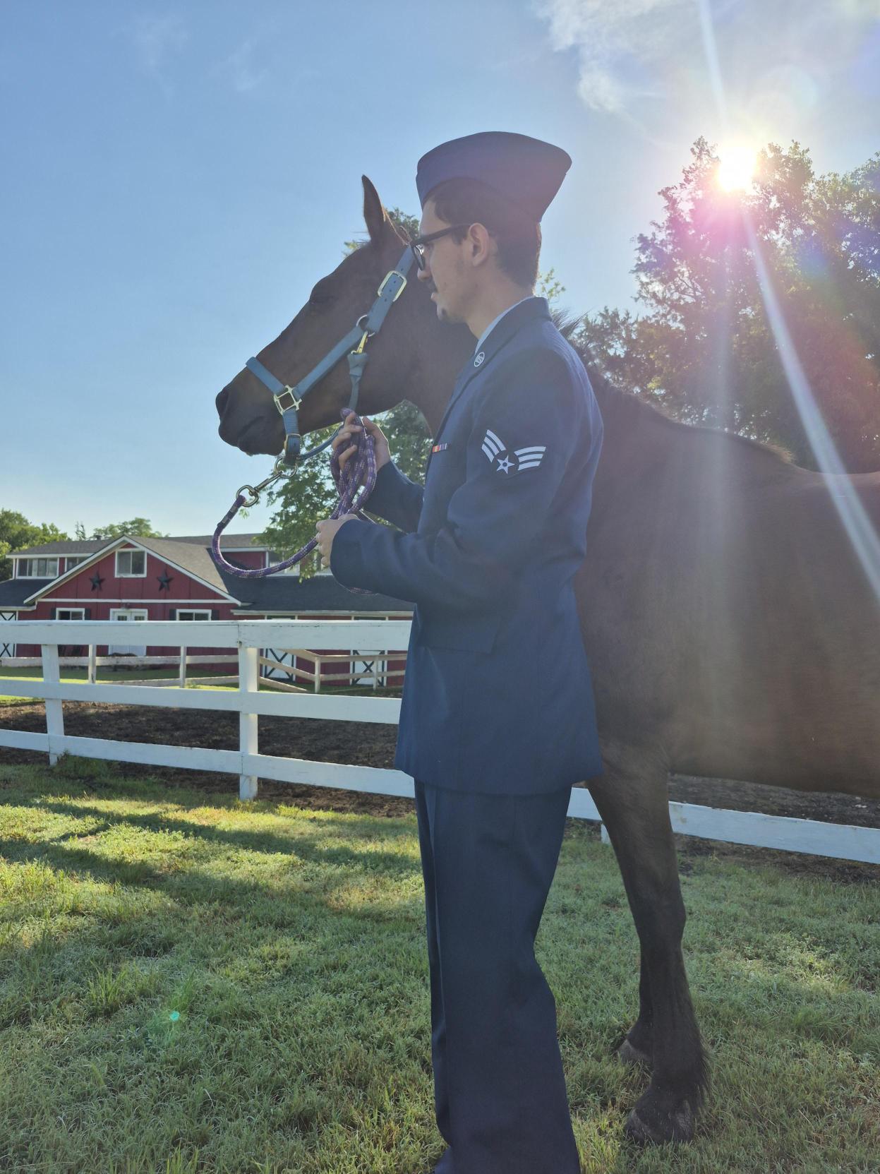 Person in military uniform holding a horse's bridle outdoors with a white fence and red building in background, sunlight shining through trees.