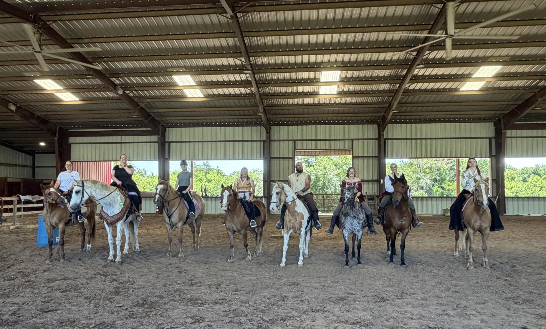 Group of people riding horses inside an indoor riding arena with a high ceiling and open sides.