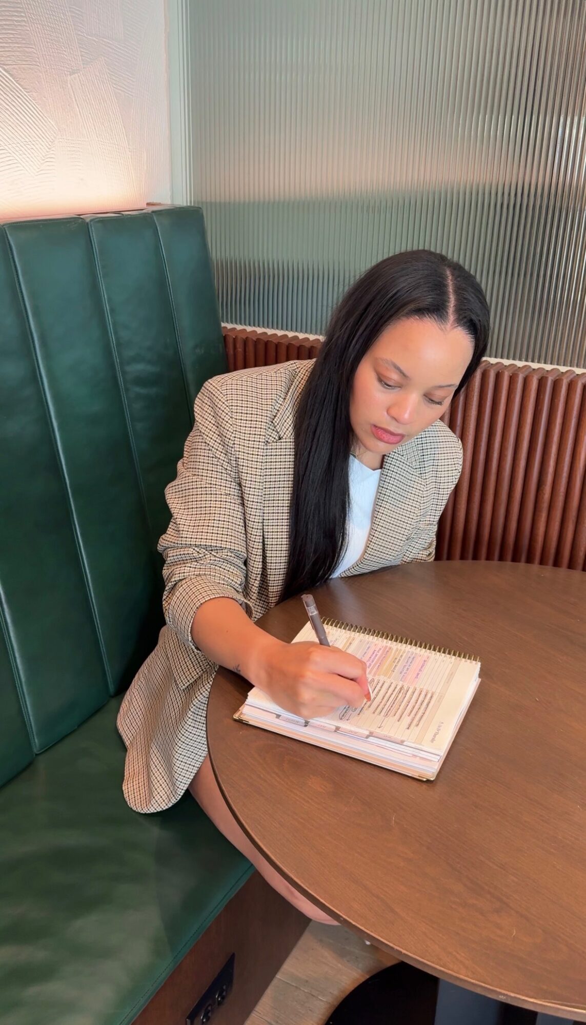 Woman with long dark hair writing in a notebook at a round table, wearing a checkered blazer, in a cafe or restaurant.