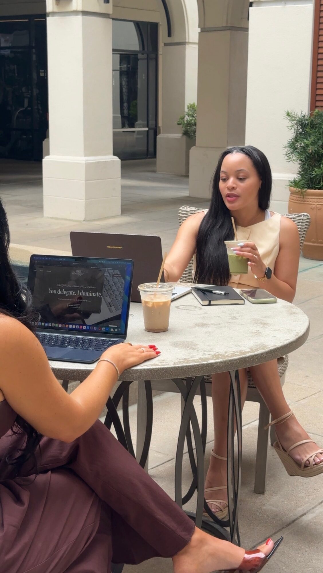 Two women sit at a round outdoor table with laptops and drinks, one using a smartphone, in a city setting.