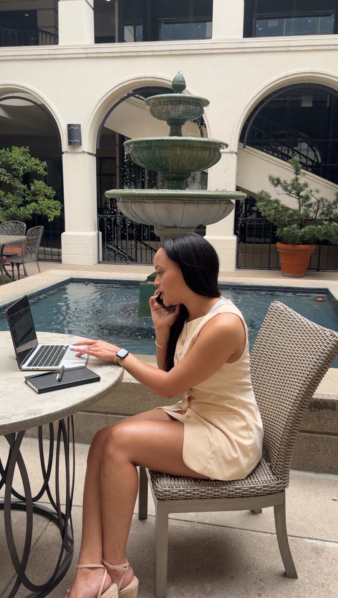 Woman sitting at outdoor table with laptop, talking on phone, fountain and building in background.